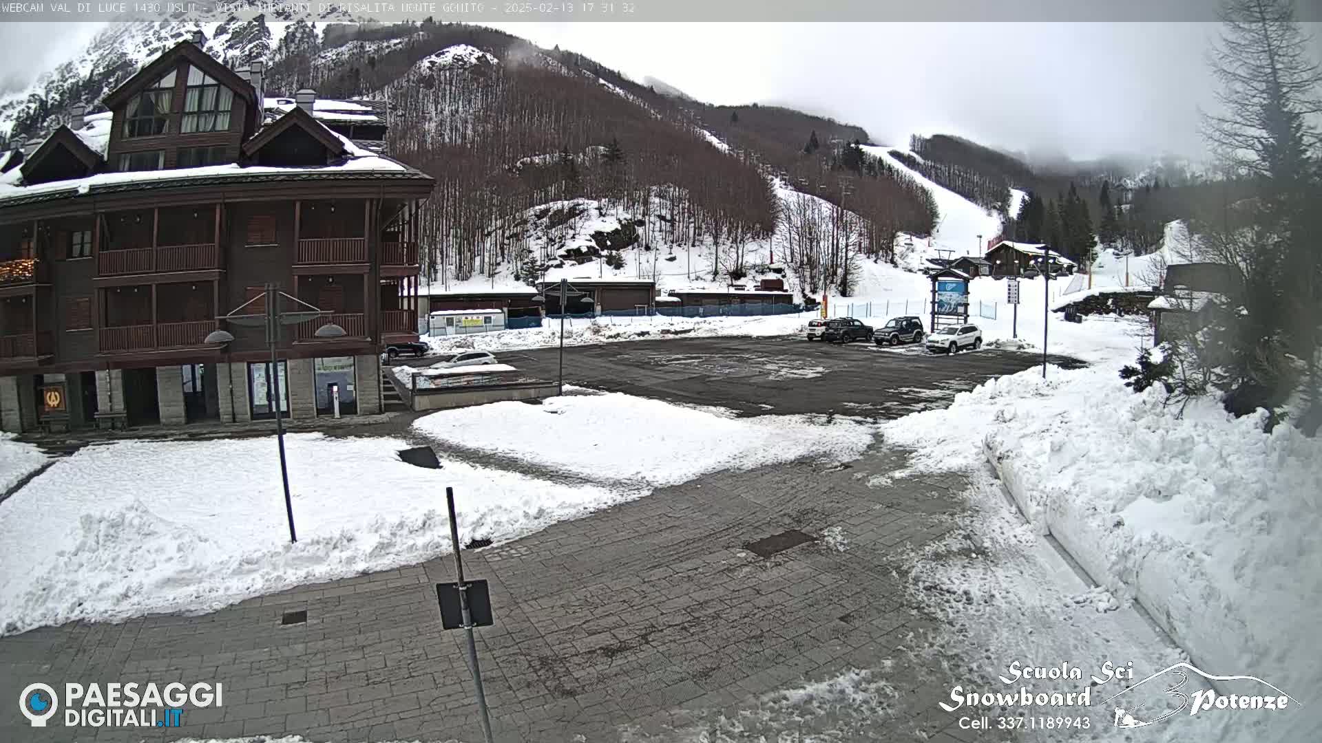 A snow-covered parking lot with several vehicles and a multi-story building is seen in the foreground, with a snow-covered mountain and ski slopes in the background under an overcast sky.