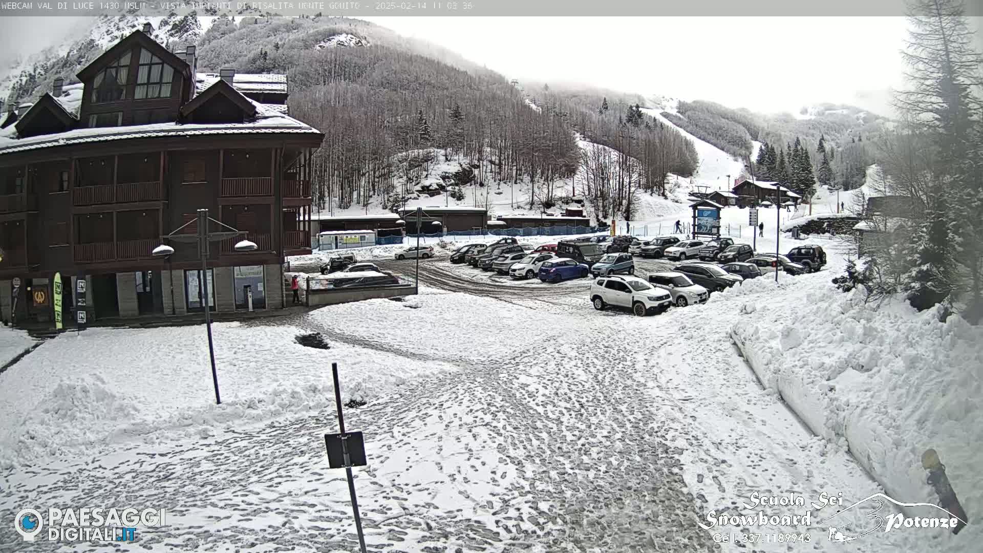 A snow-covered parking lot with numerous cars is situated in front of a multi-story brown building, with a background of a snow-dusted mountain slope featuring ski runs.