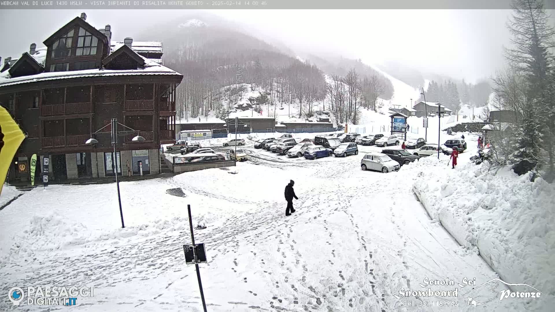 A snow-covered parking lot in front of a multi-story building, with a person walking in the snow and mountains visible in the background under a foggy sky.