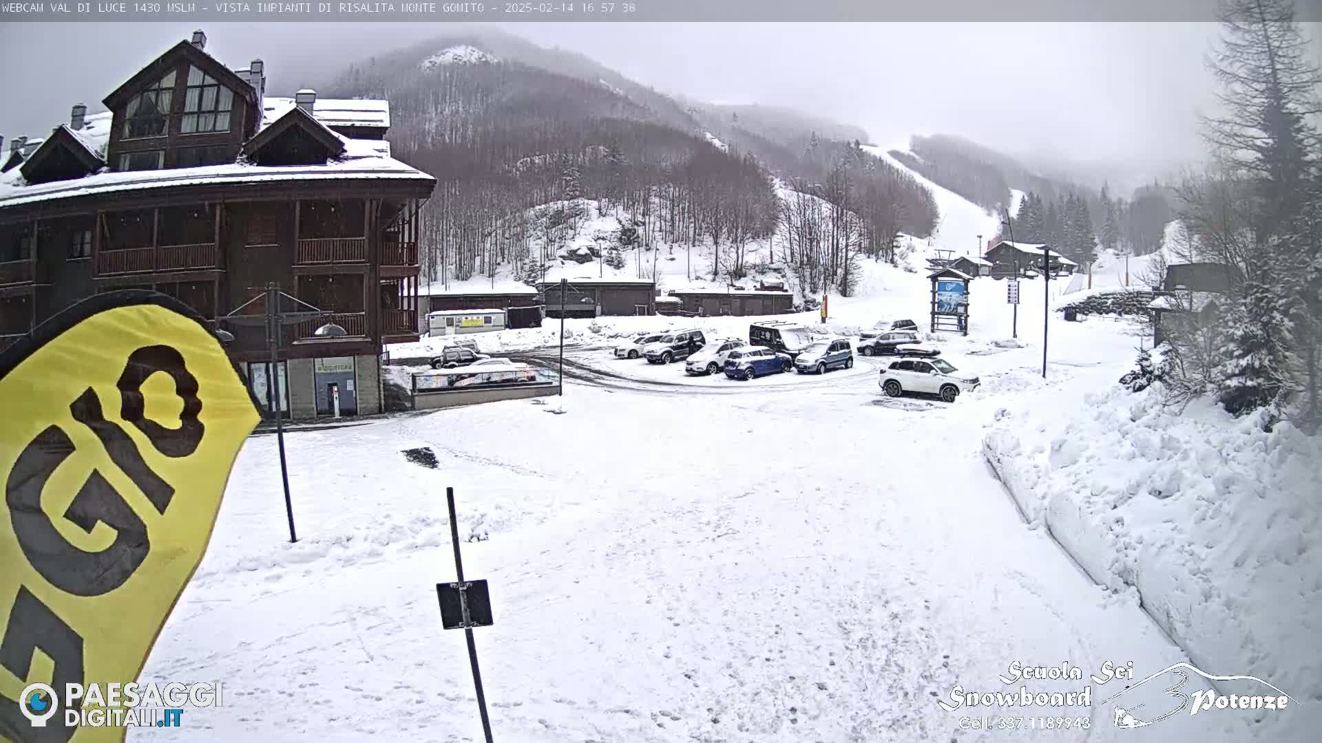 A snow-covered parking lot with several cars is in front of a multi-story building, with a snow-covered mountain and ski slope visible in the background under an overcast sky.