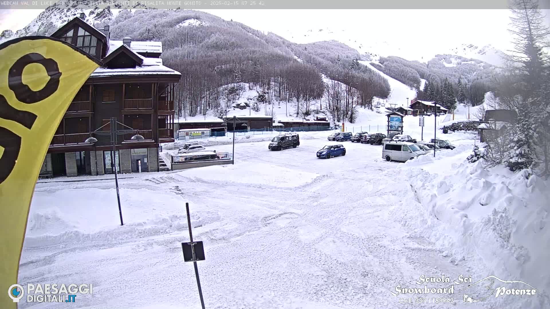 A snow-covered parking lot with several vehicles is situated in front of a multi-story building, with a snow-covered mountain and ski slope in the background.