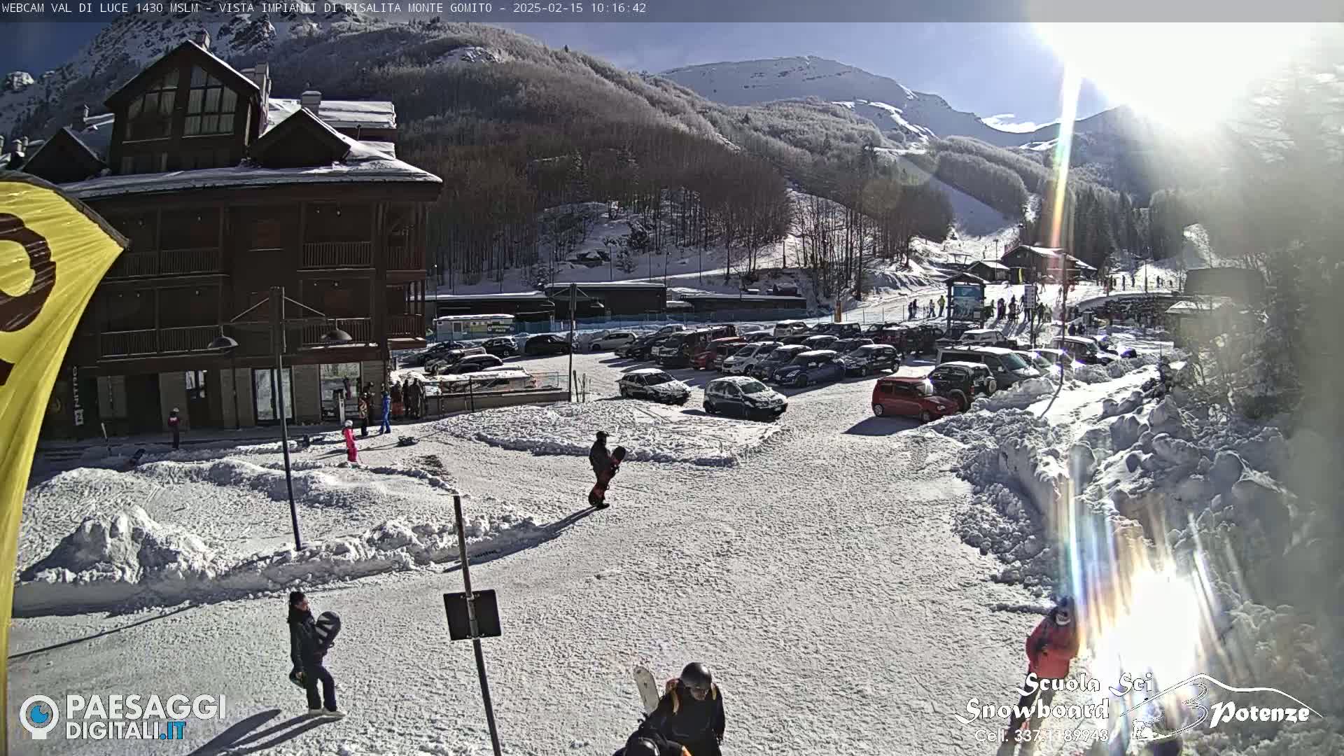 A sunny, snow-covered mountain resort with a large parking lot full of cars, a multi-story building, and several people walking around.