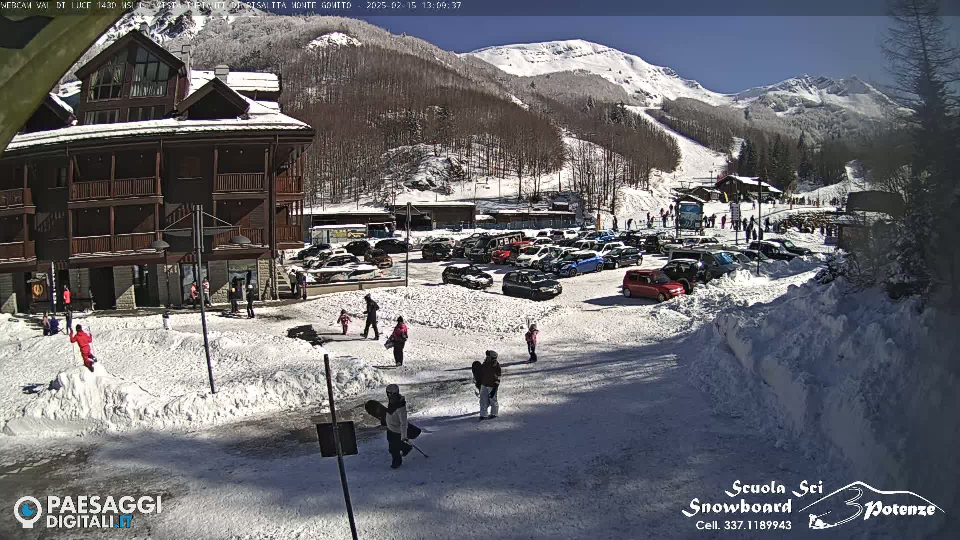 A snow-covered parking lot with a multi-story brown building and people walking around, all under a sunny sky with snow-capped mountains in the background.