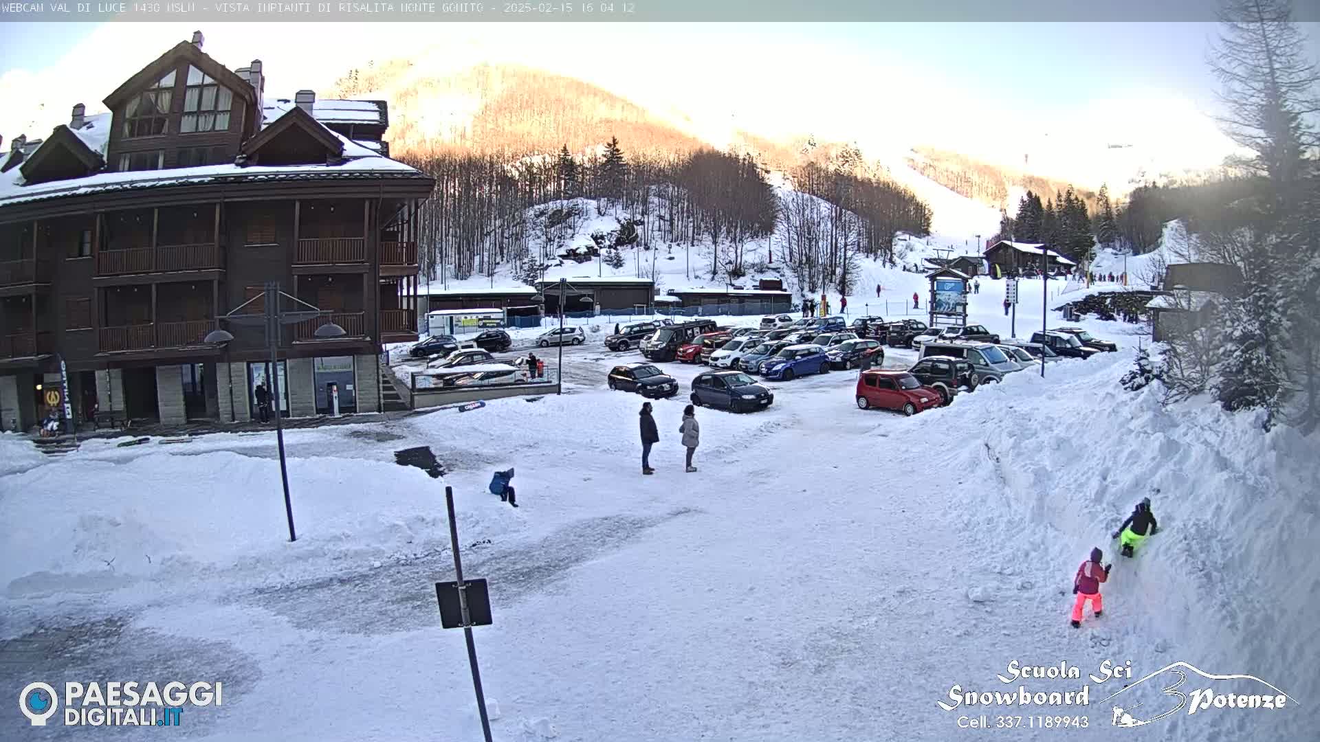 A snow-covered parking lot with several cars in front of a multi-story building, with a sunny, snowy mountainside in the background.