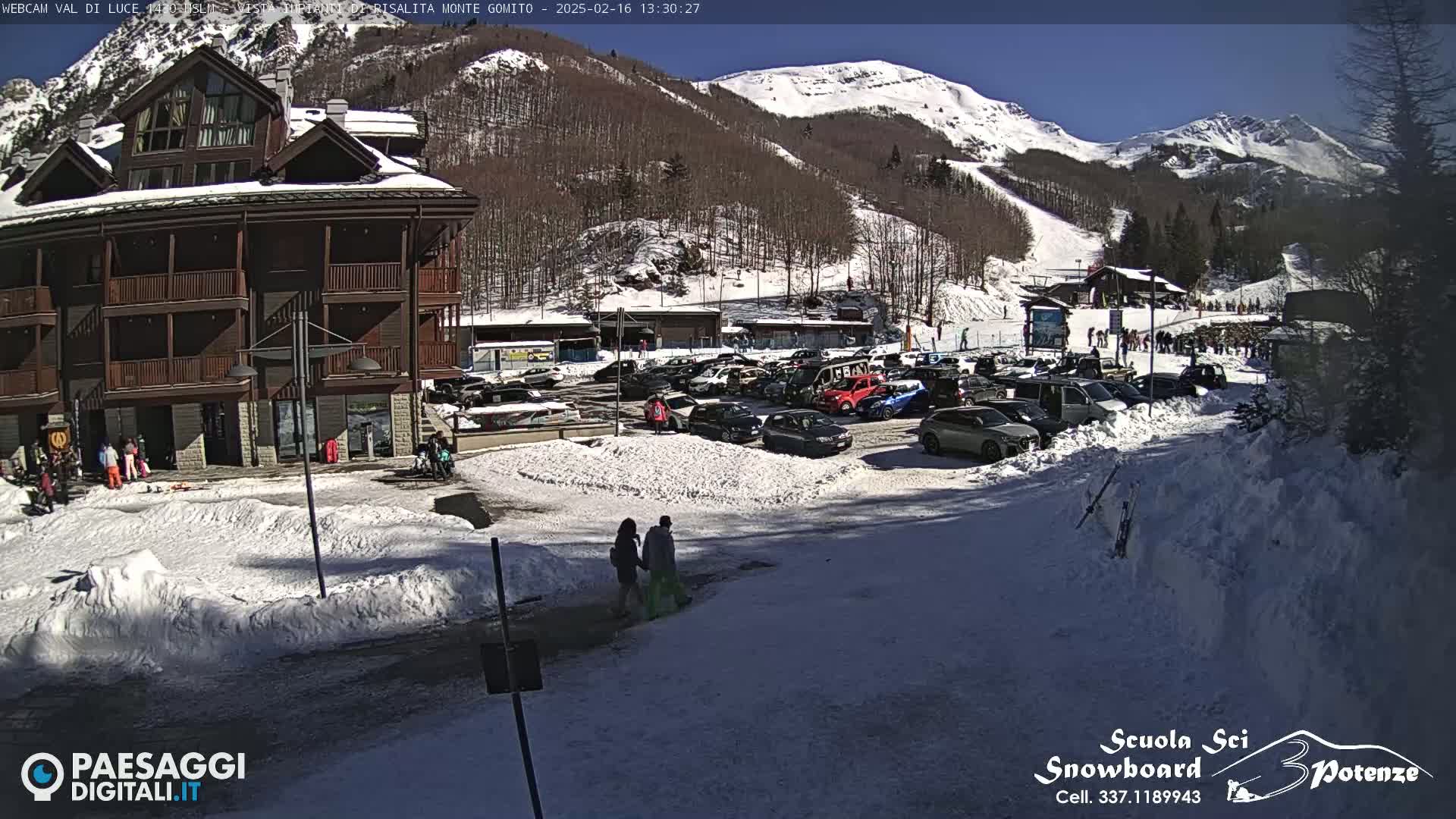 A multi-story brown building sits near a snowy parking lot filled with cars, with a sunny, snow-covered mountain in the background.