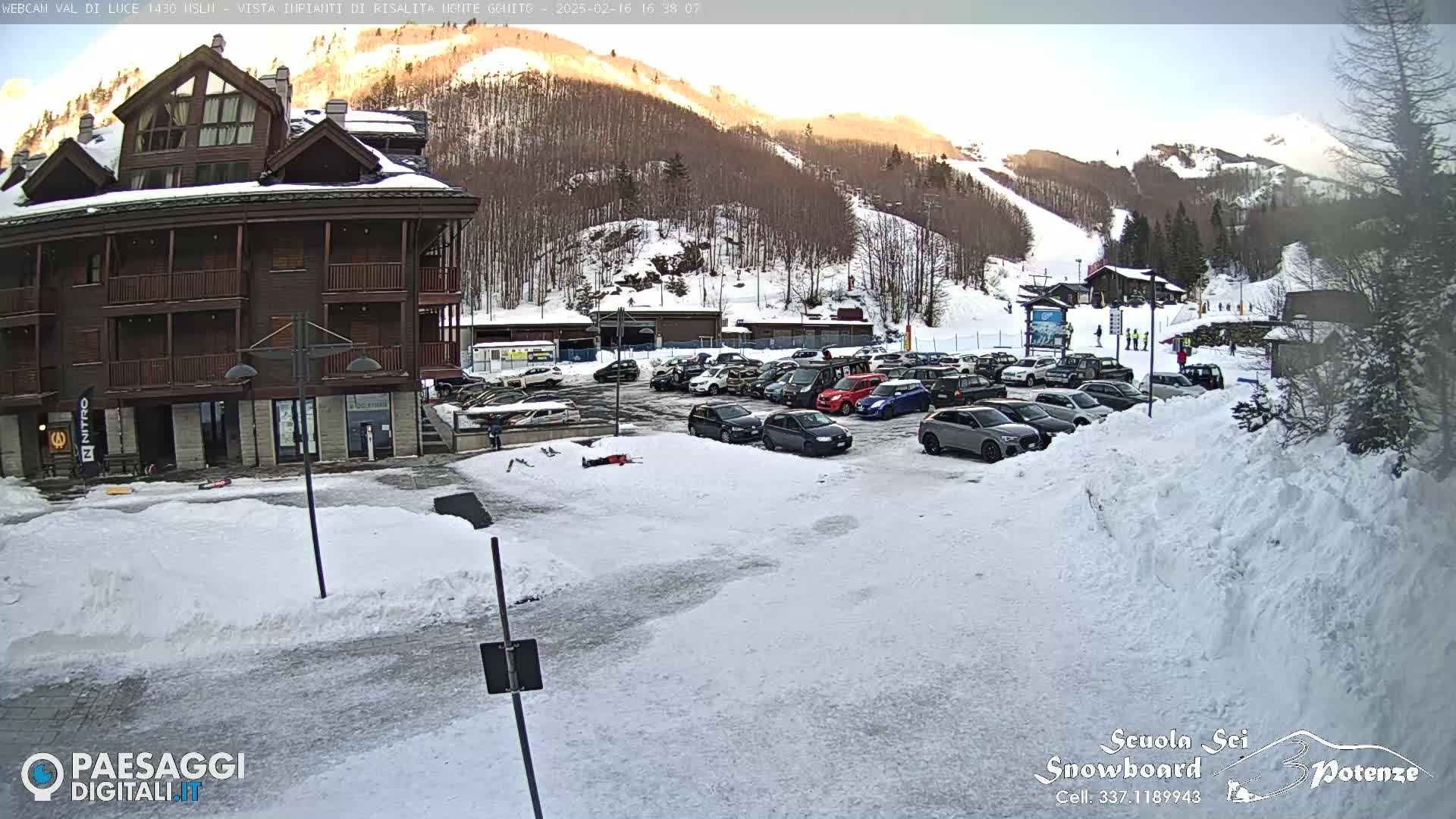 A snow-covered parking lot with cars sits in front of a brown multi-story building, with snow-covered mountains and ski slopes in the background under a partly sunny sky.