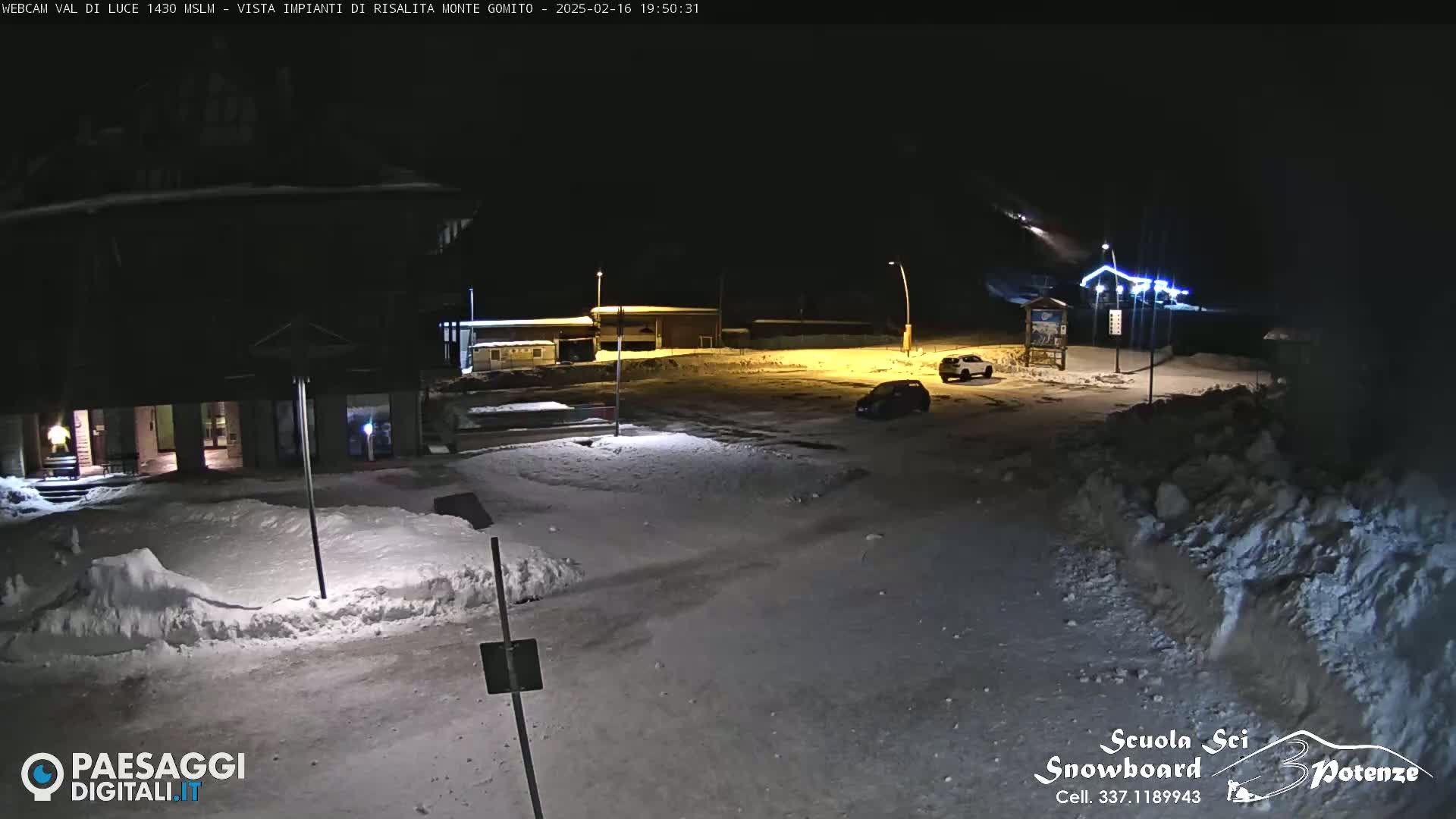 A snow-covered parking lot at night, illuminated by artificial lights, with a few cars parked and buildings in the background.
