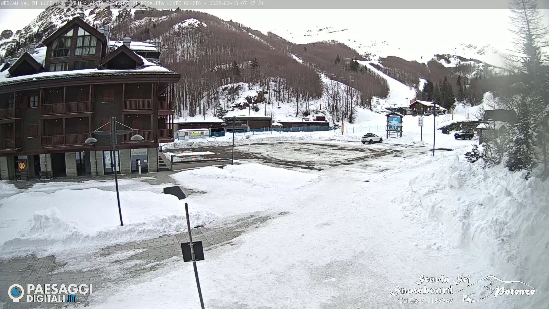 A snow-covered parking lot sits in front of a multi-story brown building, with a snowy mountain and ski slopes in the background.