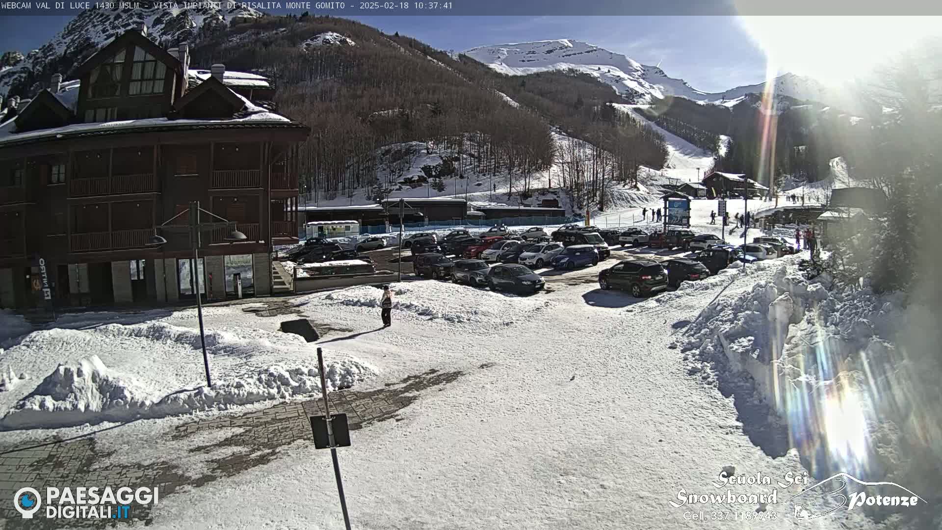 A sunny, snow-covered mountainside parking lot with a multi-story building and numerous cars parked, near ski slopes.