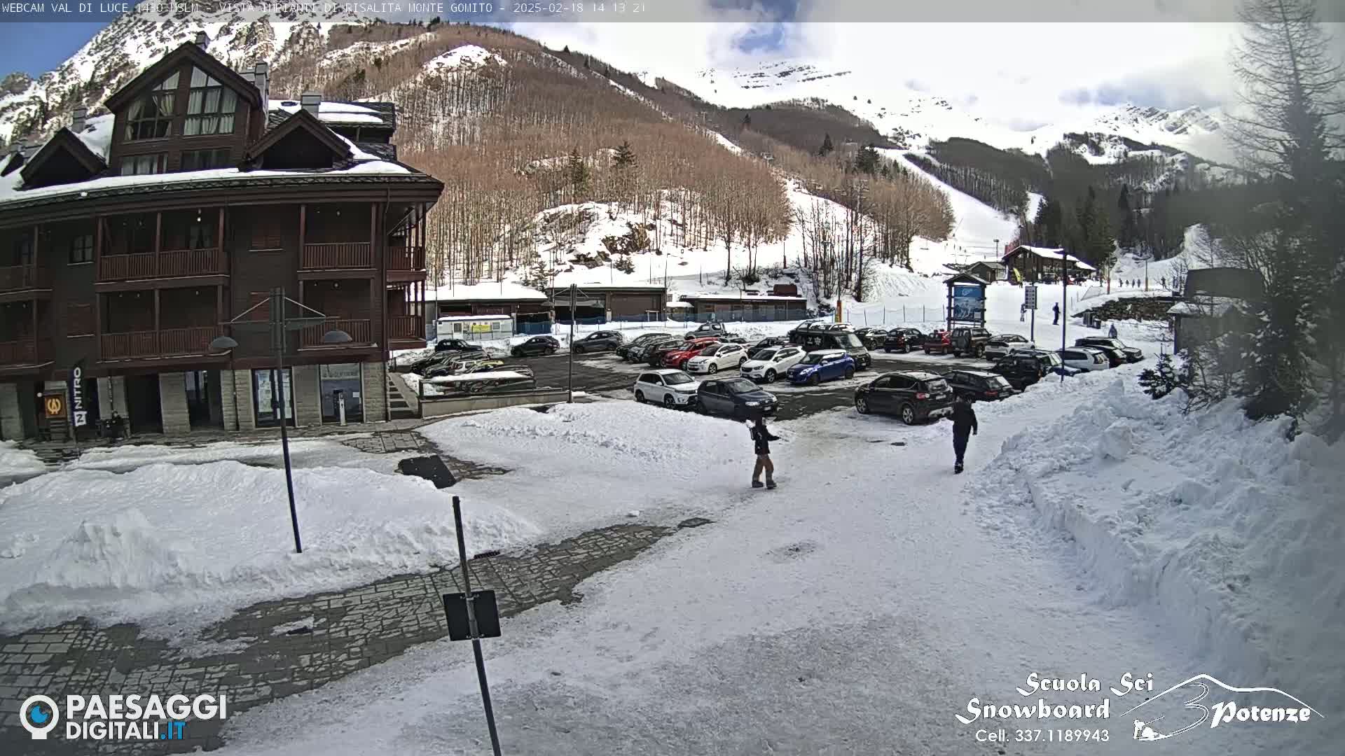 A snow-covered parking lot with cars and people is situated in front of a multi-story brown building, with a snow-covered mountain and ski slopes in the background under a partly cloudy sky.