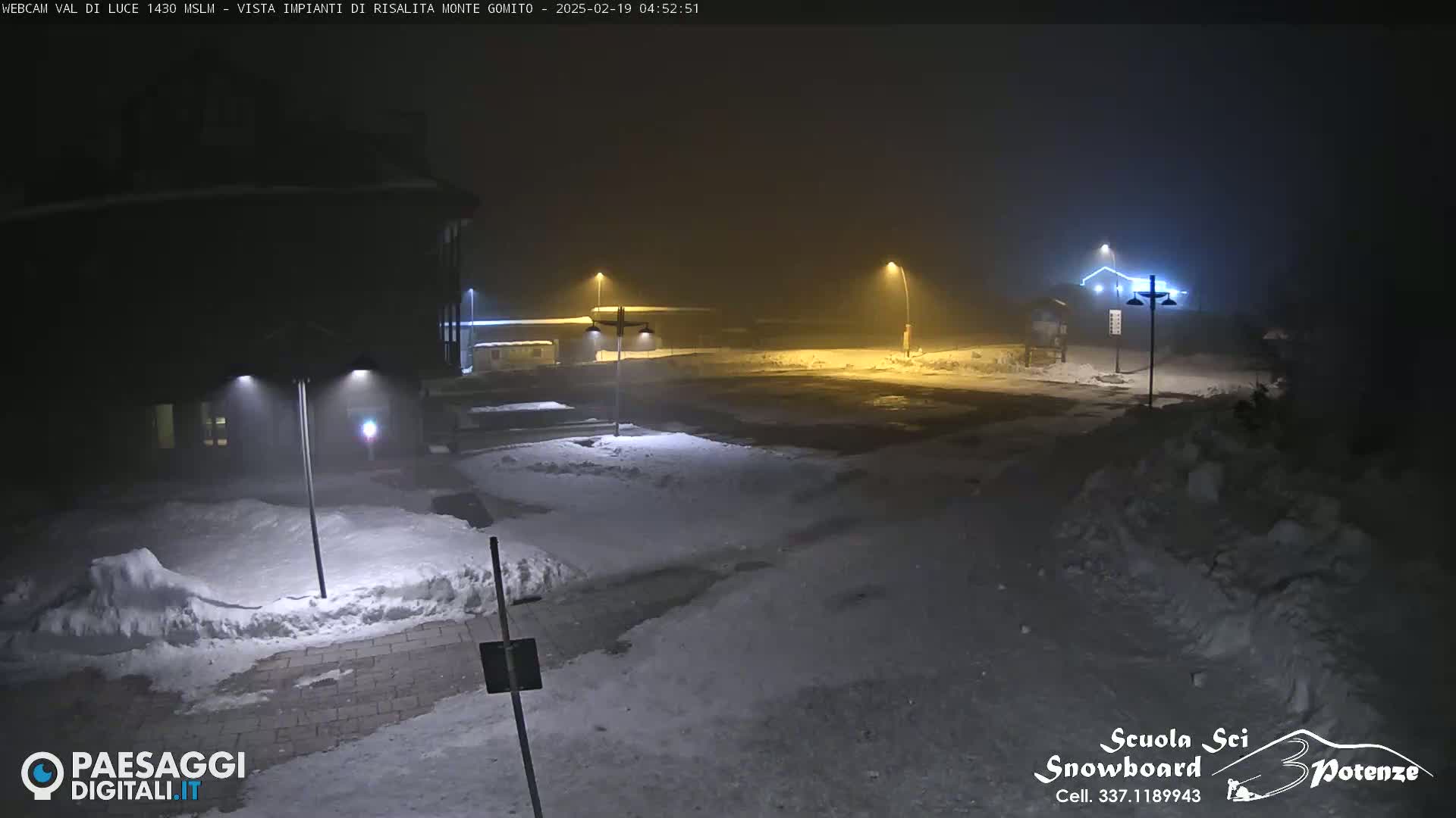 A snow-covered parking lot and building are dimly lit by streetlights in foggy nighttime conditions.