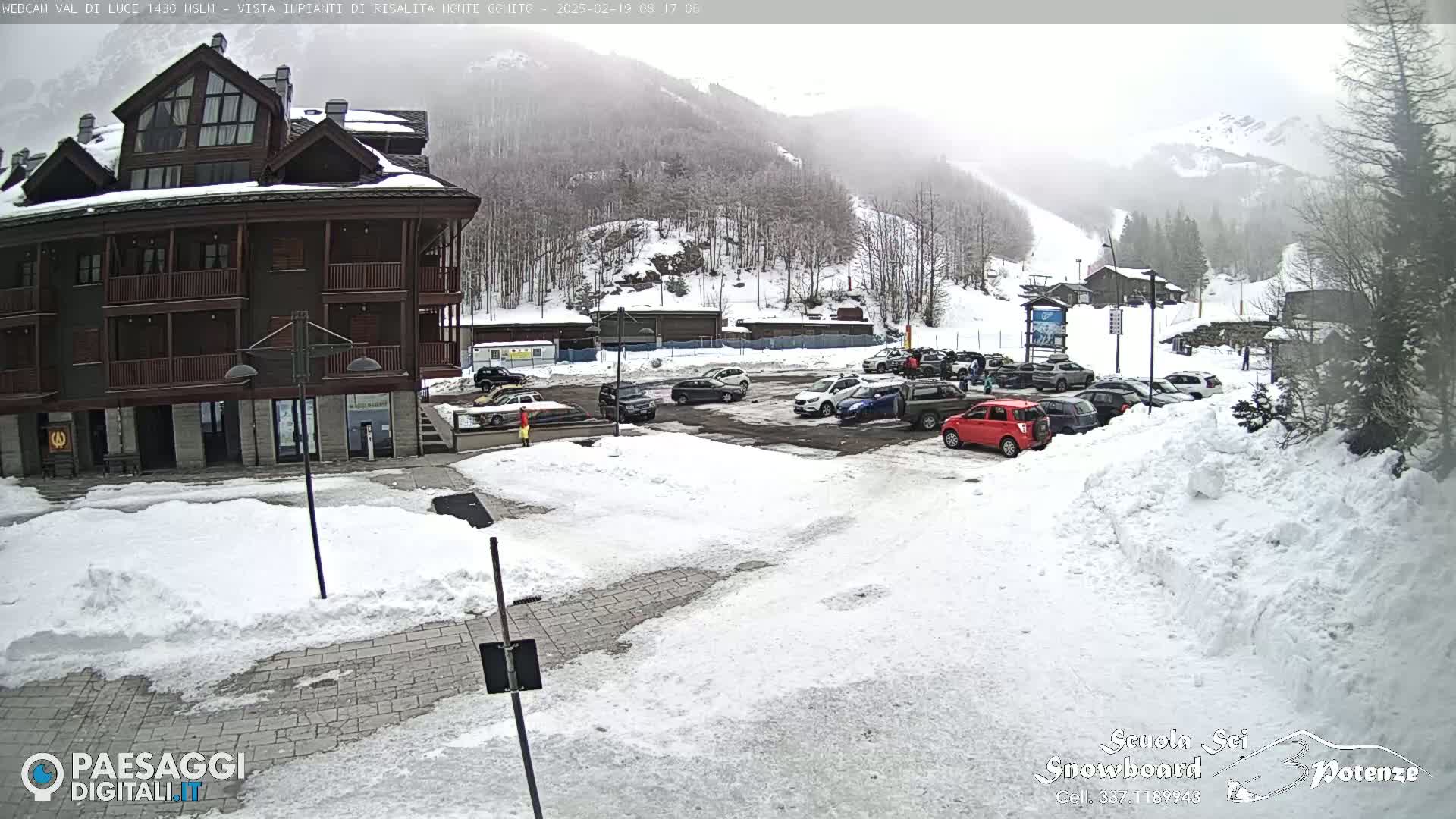 A snow-covered parking lot with several cars is in front of a multi-story brown building, with a snowy mountainside and ski slope visible in the background under an overcast sky.