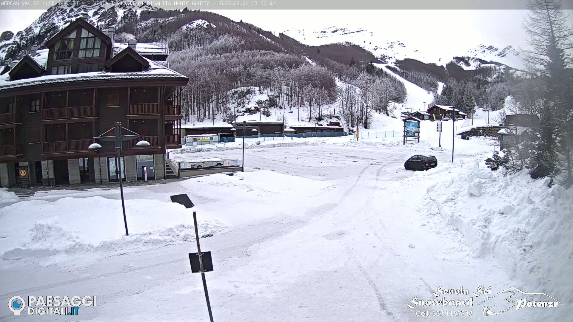 A snow-covered parking lot in front of a brown multi-story building, with a car parked and a snowy mountainside in the background.