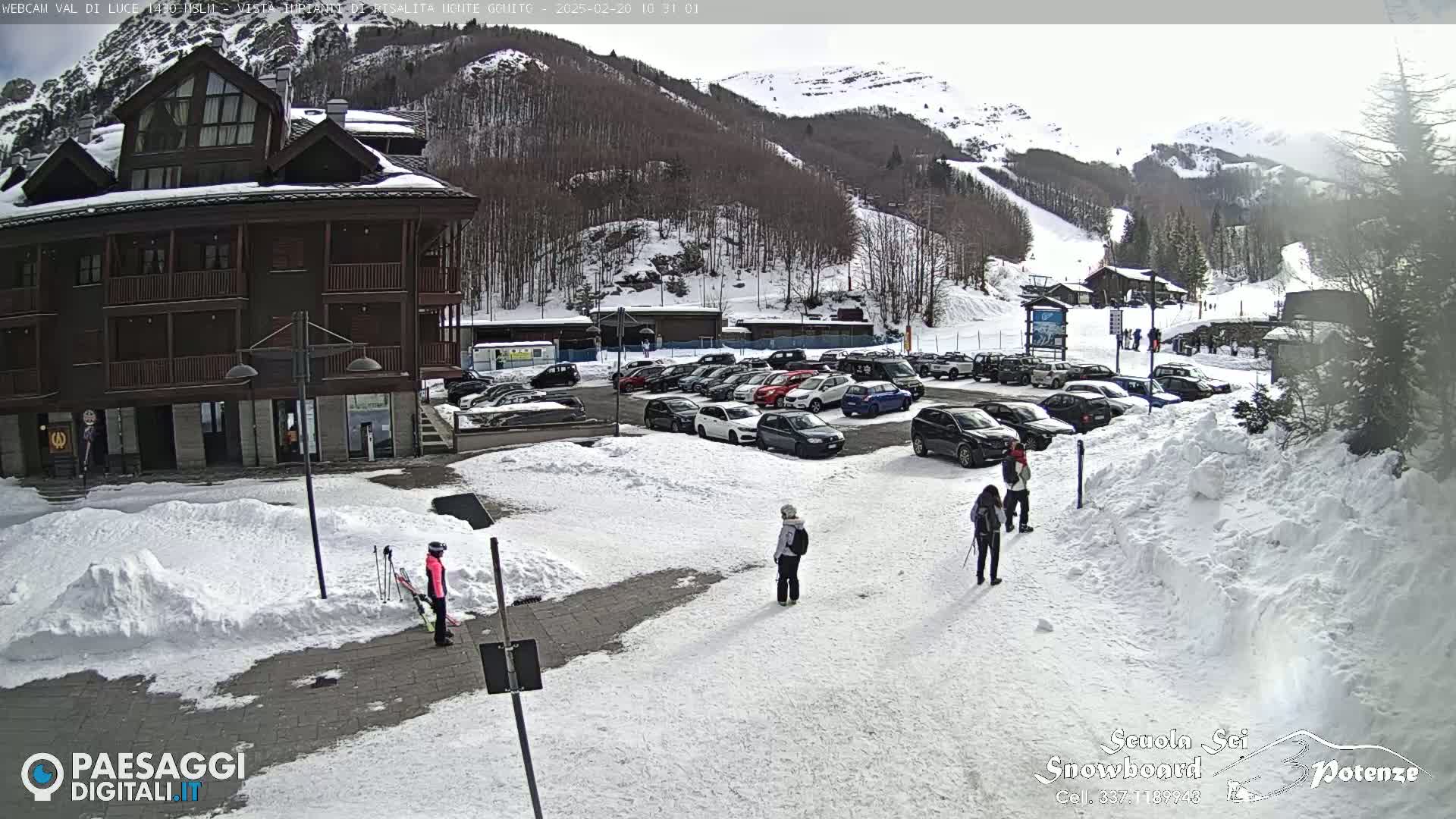 A snow-covered parking lot with several cars and a few people is situated in front of a multi-story brown building, with a ski slope and snowy mountains visible in the background under a bright, mostly clear sky.