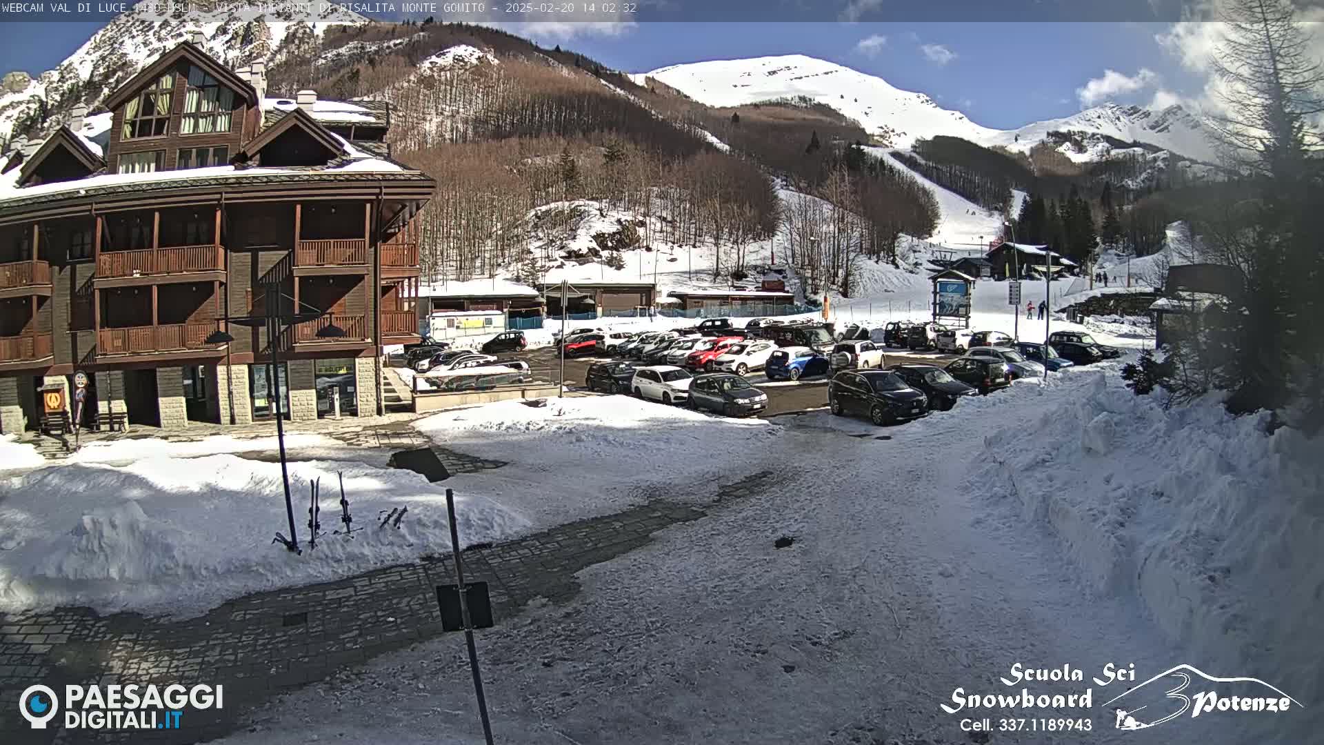 A snow-covered parking lot with cars sits in front of a multi-story building, all nestled at the base of a sunny, snow-capped mountain.