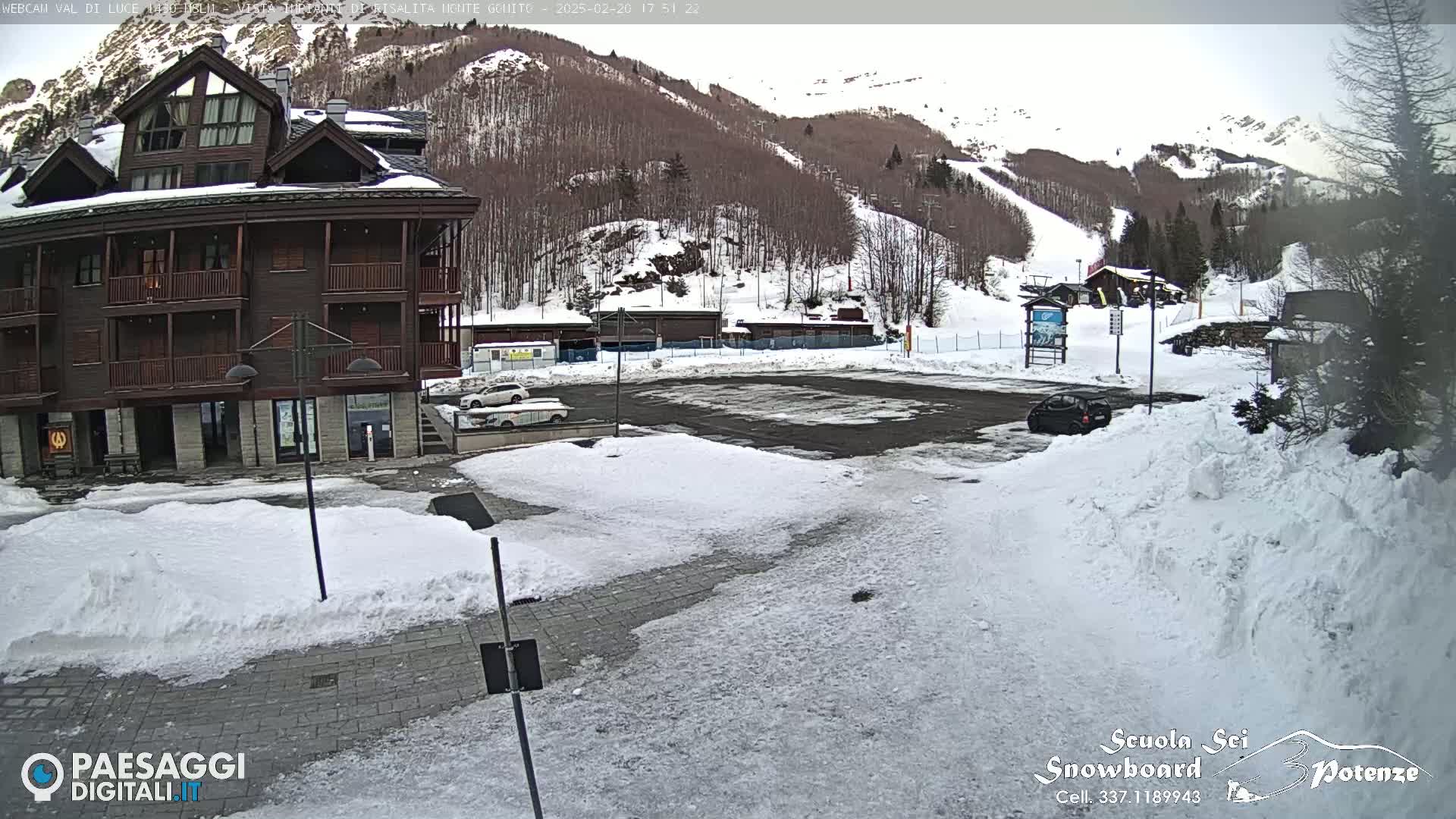 A snow-covered parking lot sits in front of a dark brown multi-story building, with a mountainside visible in the background under a cloudy sky.