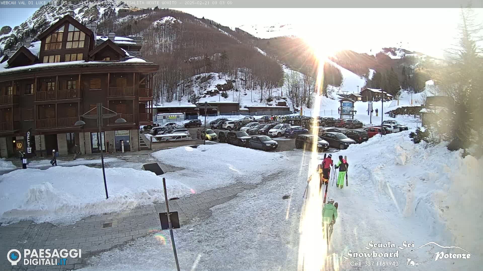 A snow-covered parking lot with several cars and a few skiers walking by a multi-story building, all under a bright, sunny winter sky.