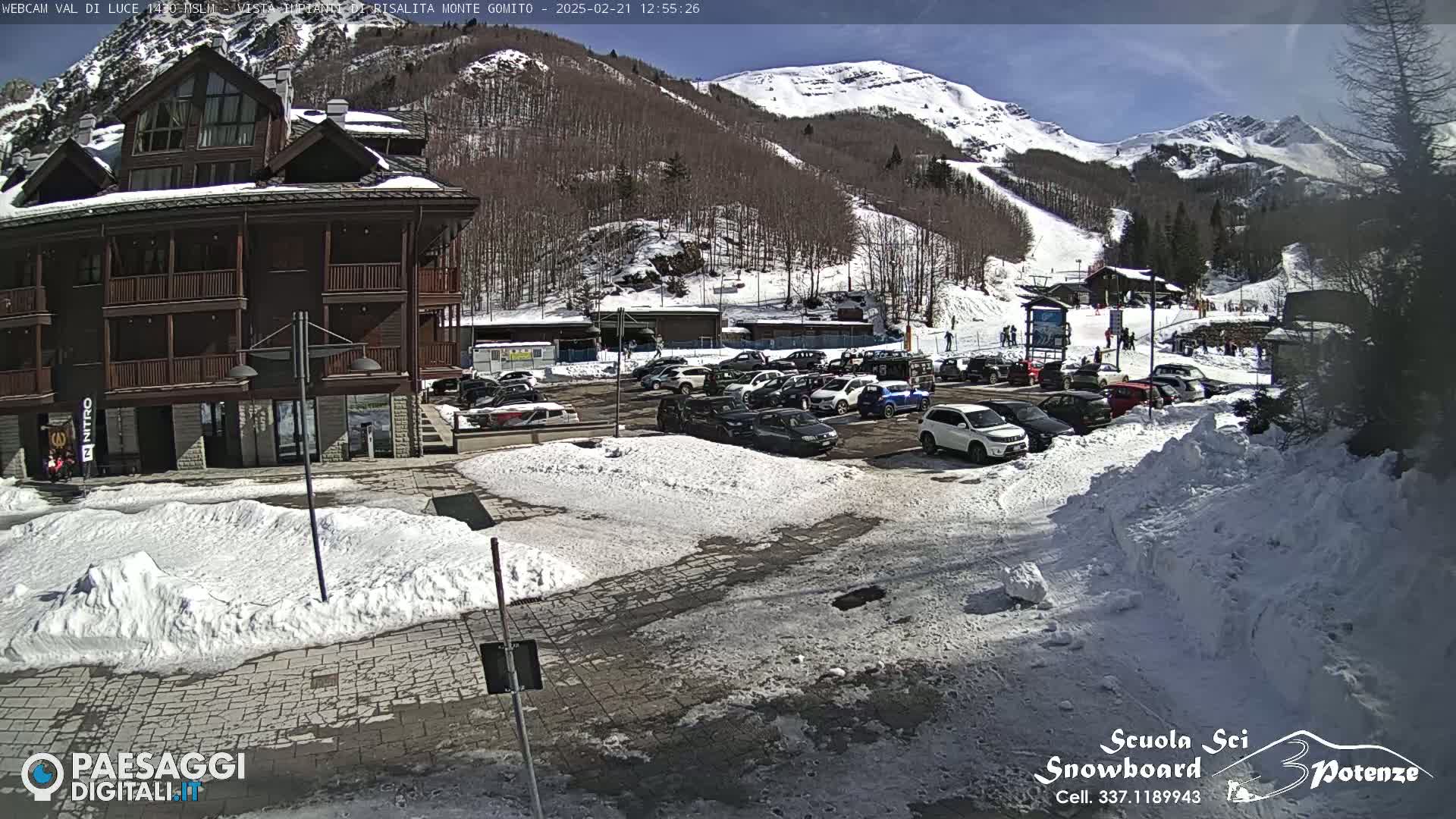 A sunny day in a snow-covered mountain town shows a parking lot full of cars in front of a multi-story brown building and a ski slope in the background.