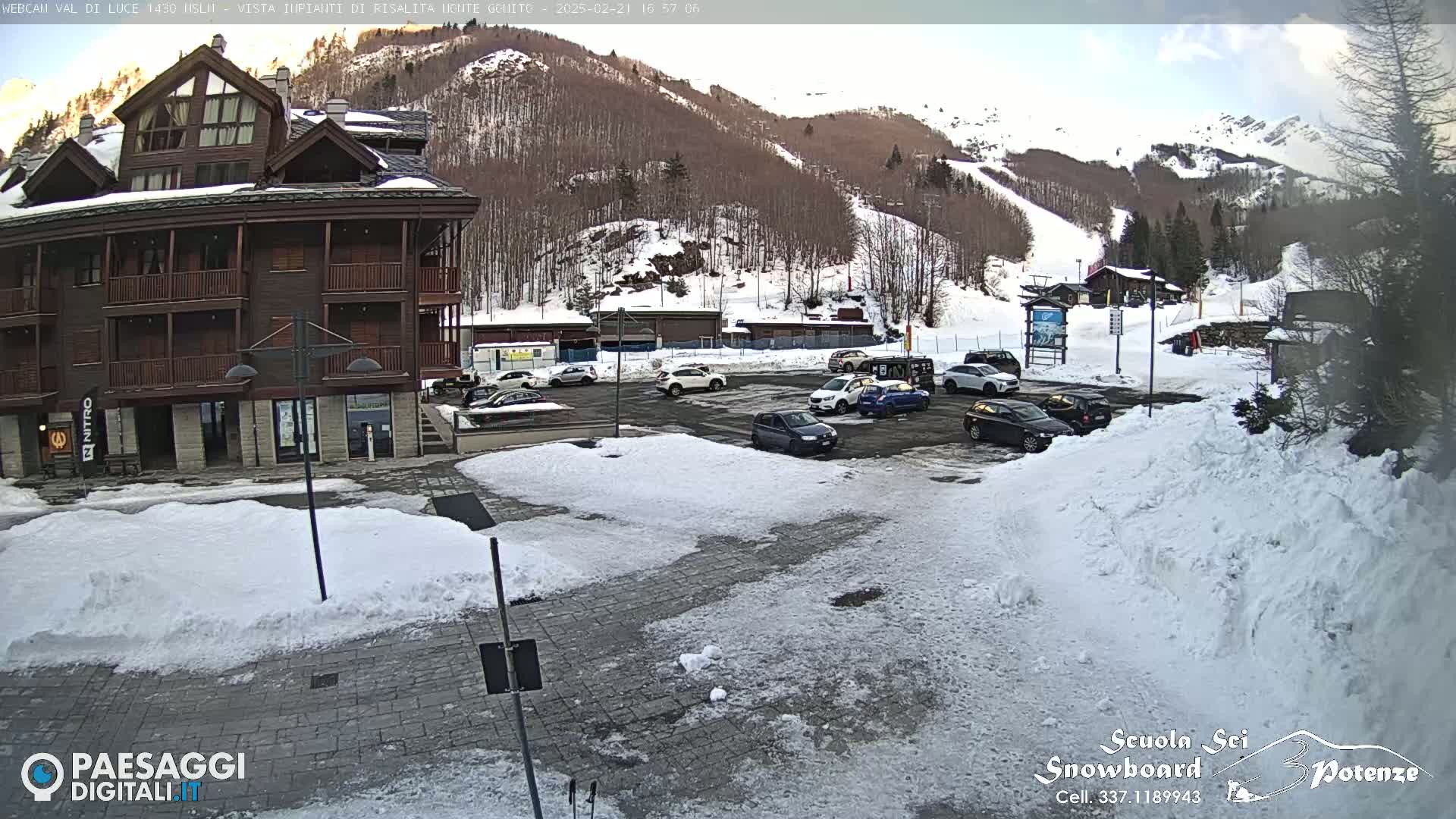 A partially snow-covered parking lot sits in front of a multi-story brown building, with a snow-covered mountain and ski slopes in the background under a partly cloudy sky.