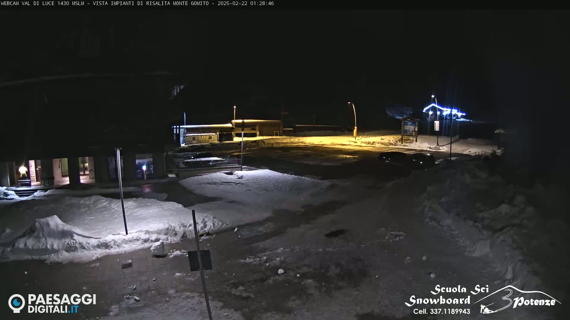 A snow-covered nighttime scene shows buildings illuminated by artificial lights, with a mostly empty parking lot and patches of snow.