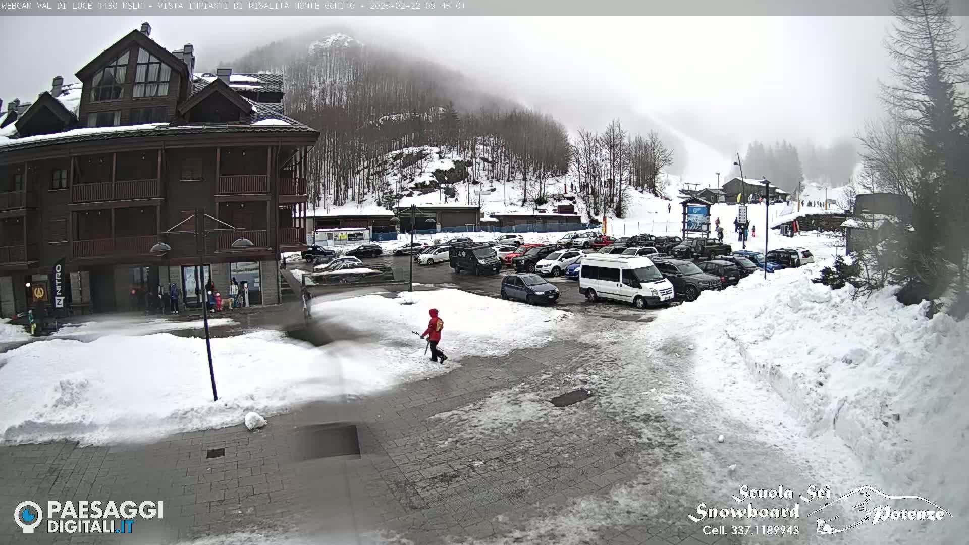 A snow-covered parking lot in front of a multi-story brown building, with a foggy mountain in the background.