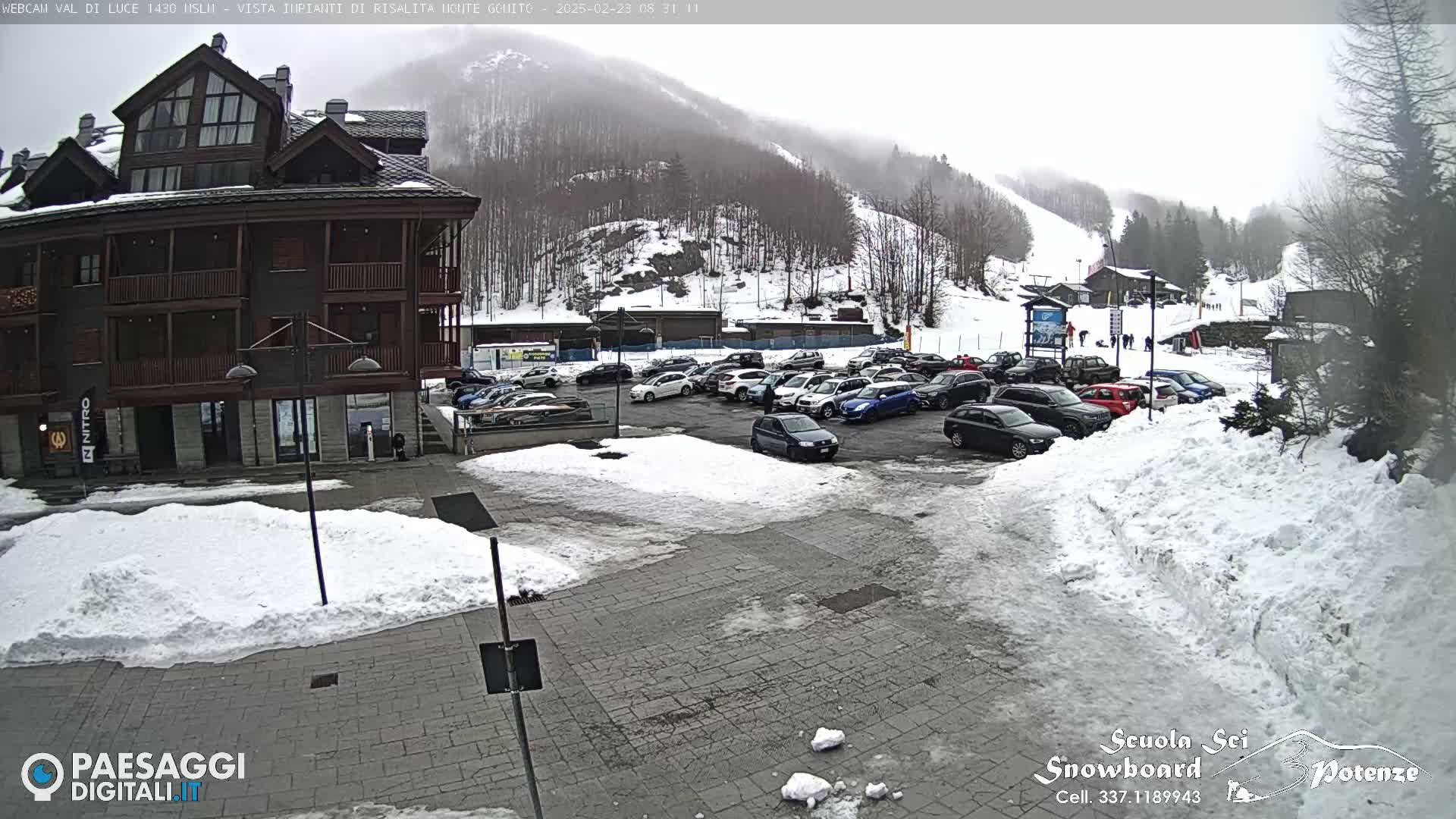A snow-covered parking lot with cars in front of a multi-story building, all situated on a mountainside in overcast weather.
