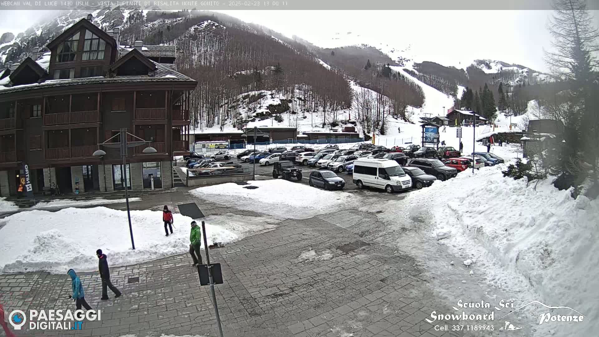 A snow-covered parking lot with several cars and a multi-story brown building sits in front of a snow-dusted mountain with a ski slope.