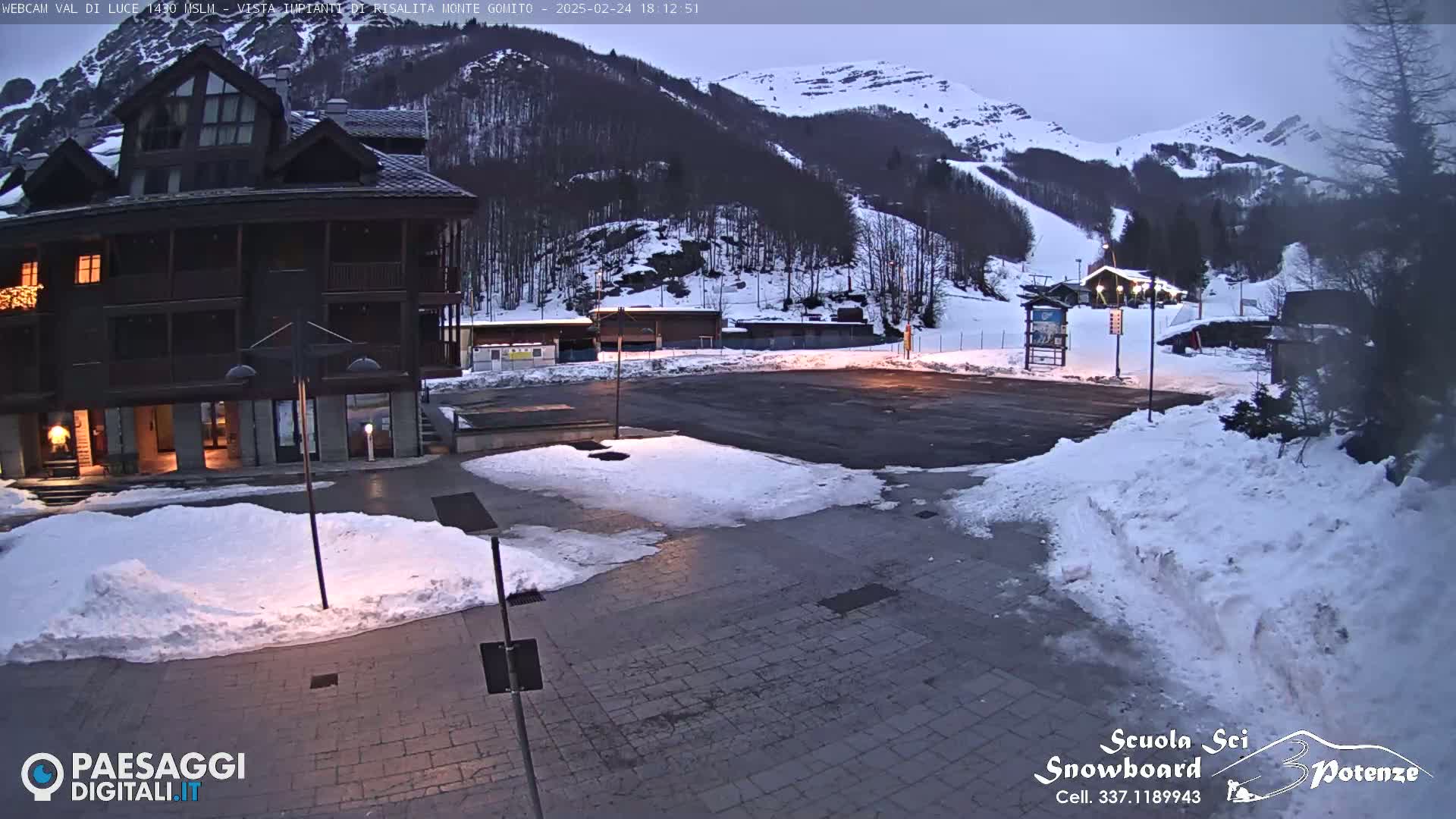 A snow-covered plaza sits before a dark-brown building, with a snow-covered mountain and ski slope in the background under a twilight sky.