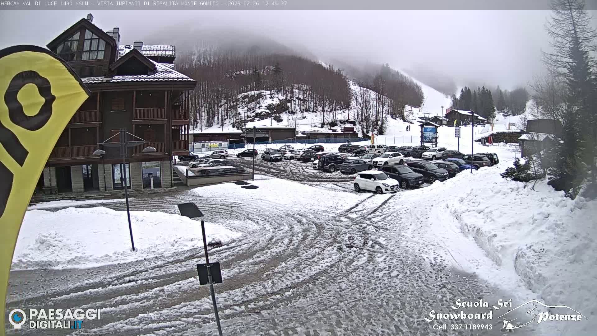 A snow-covered parking lot with cars and a multi-story building is visible, with a foggy, snowy mountainside in the background.