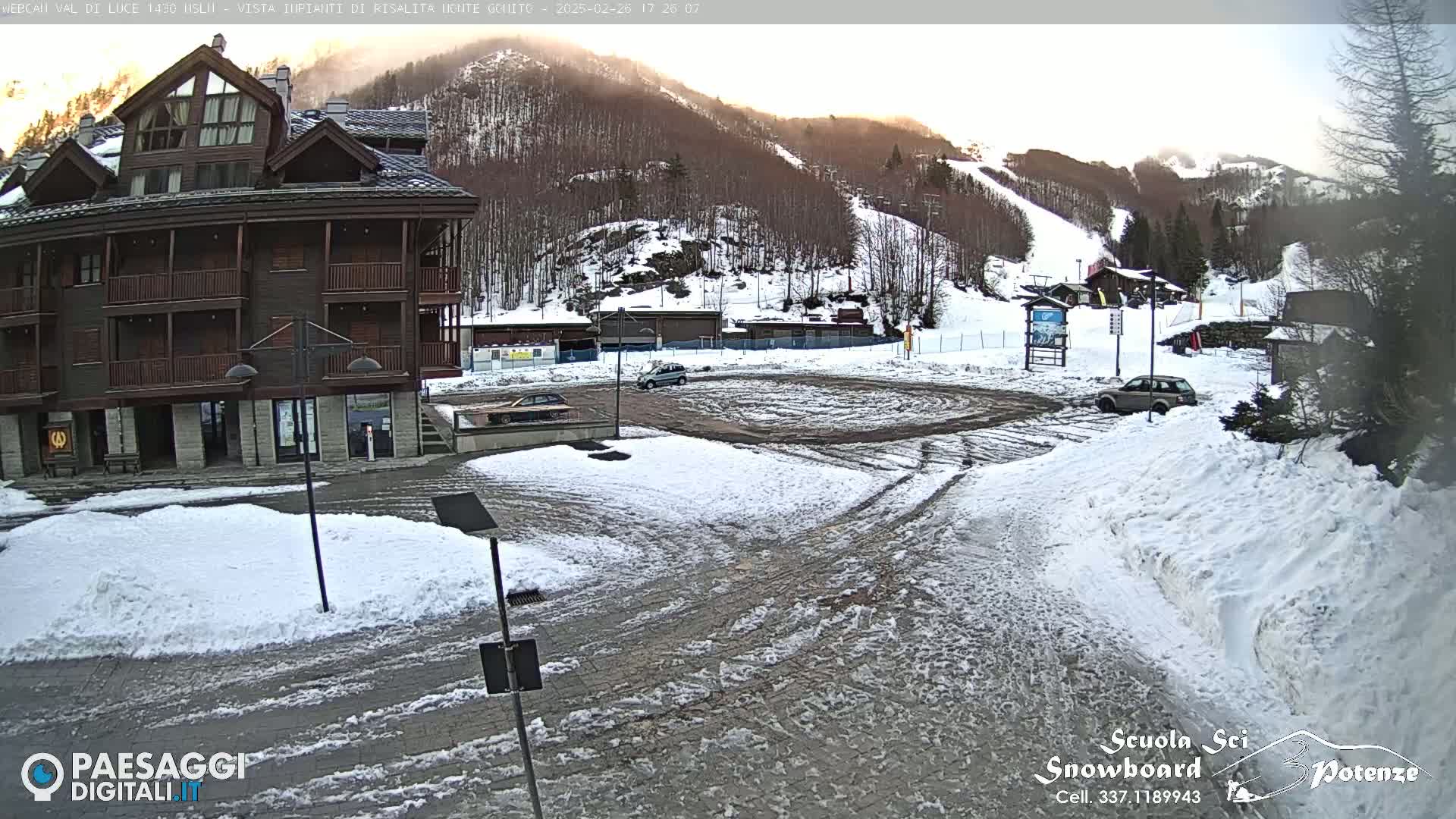 A snow-covered parking lot and a multi-story wood building sit at the base of a snow-dusted mountain with ski slopes visible in the background under a partly sunny sky.