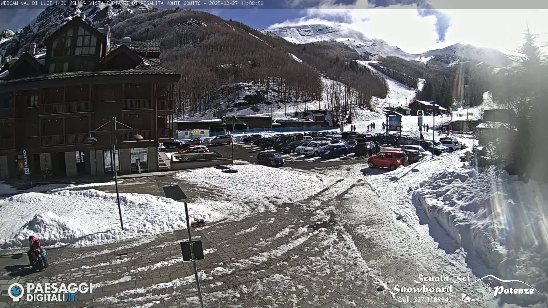 A sunny day in a snow-covered mountain resort town shows a parking lot full of cars near a multi-story building, with a ski slope visible in the background.