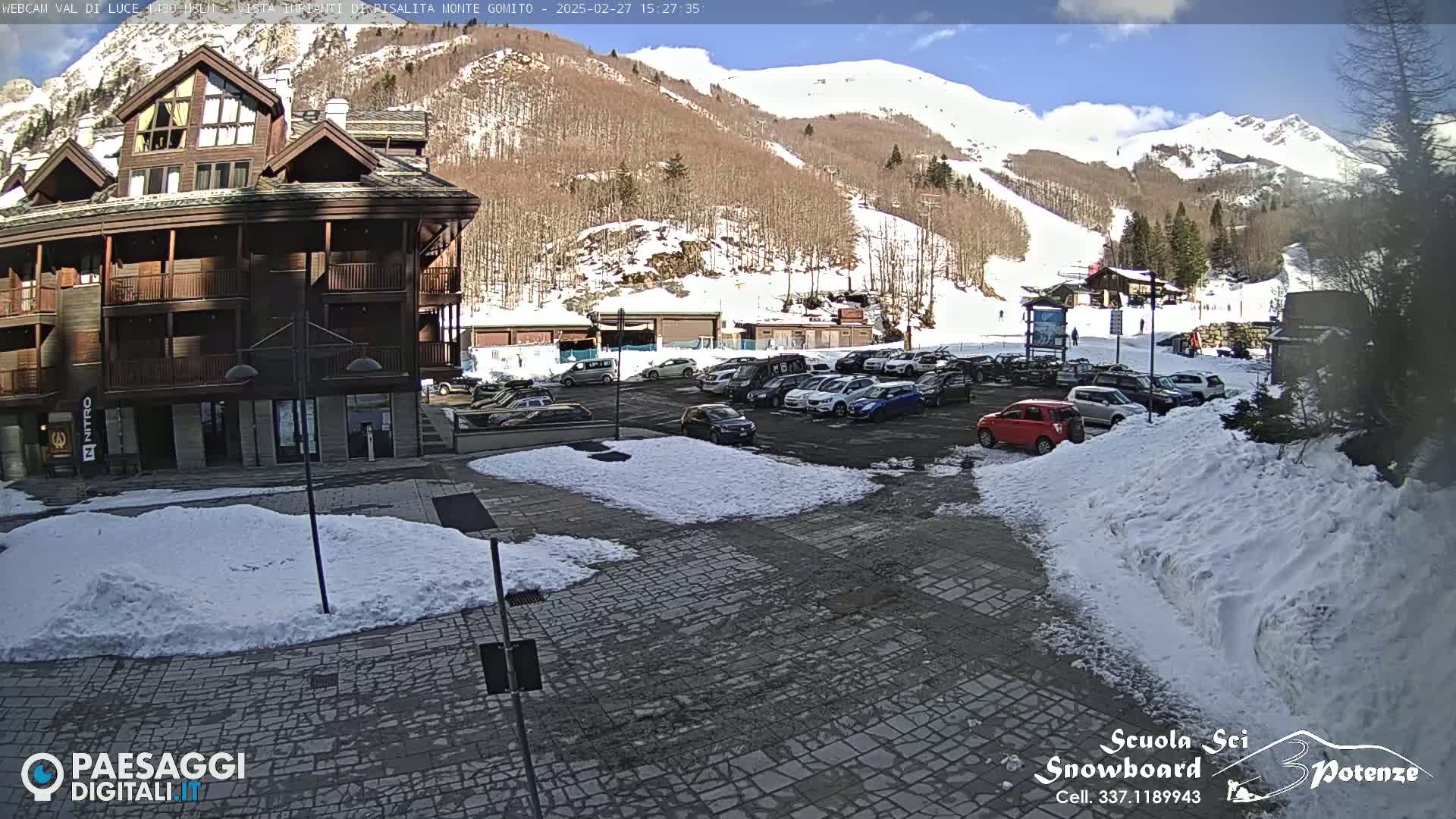 A sunny day reveals a snow-covered parking lot filled with cars in front of a multi-story building near a ski slope on a snow-dusted mountain.