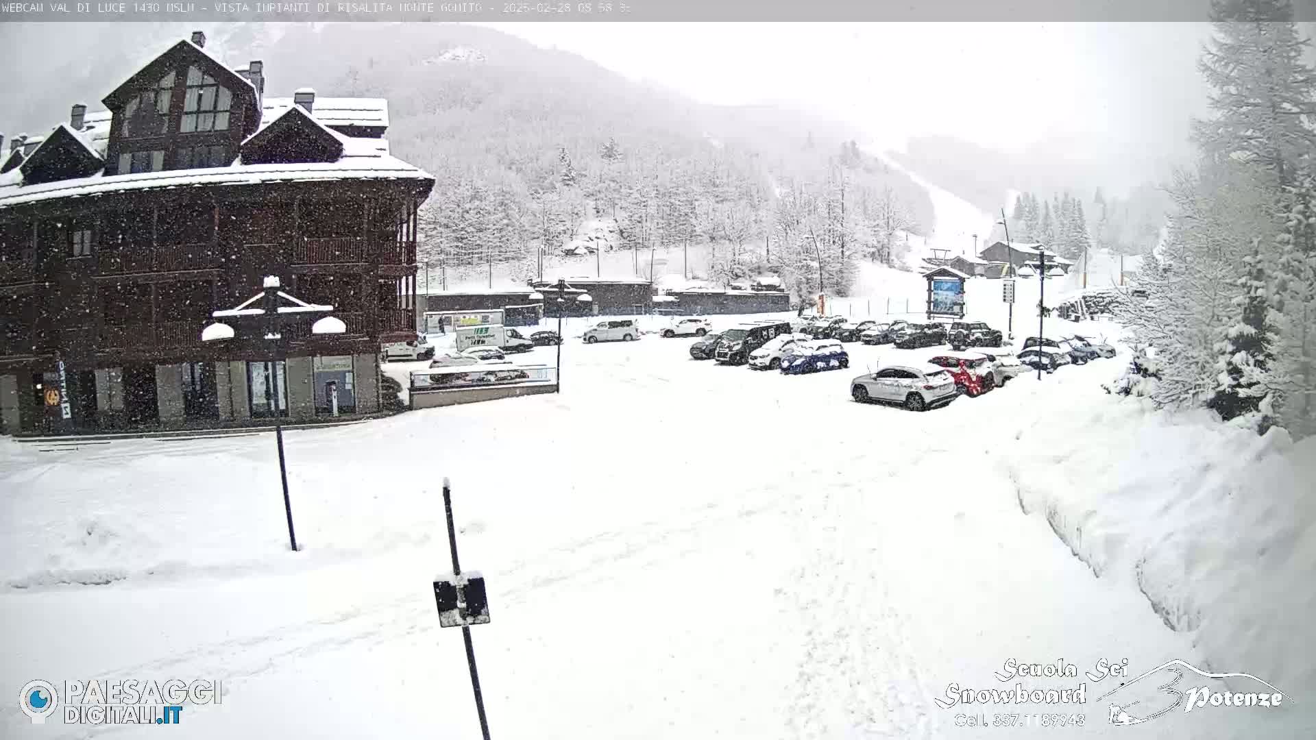 A snow-covered parking lot with cars is in front of a multi-story building, with snow falling and snow-covered mountains in the background.