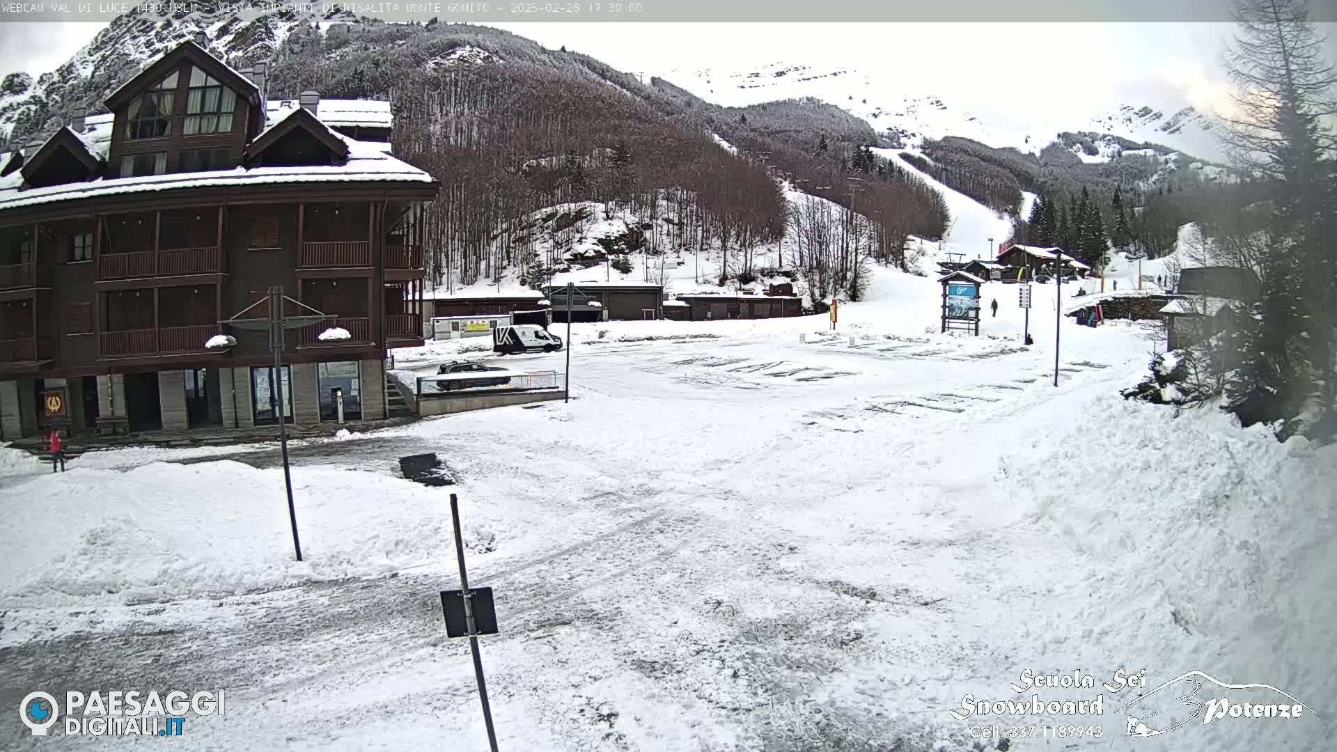 A snow-covered parking lot and a multi-story brown building are situated at the base of a snow-dusted mountain with ski slopes visible in the background.