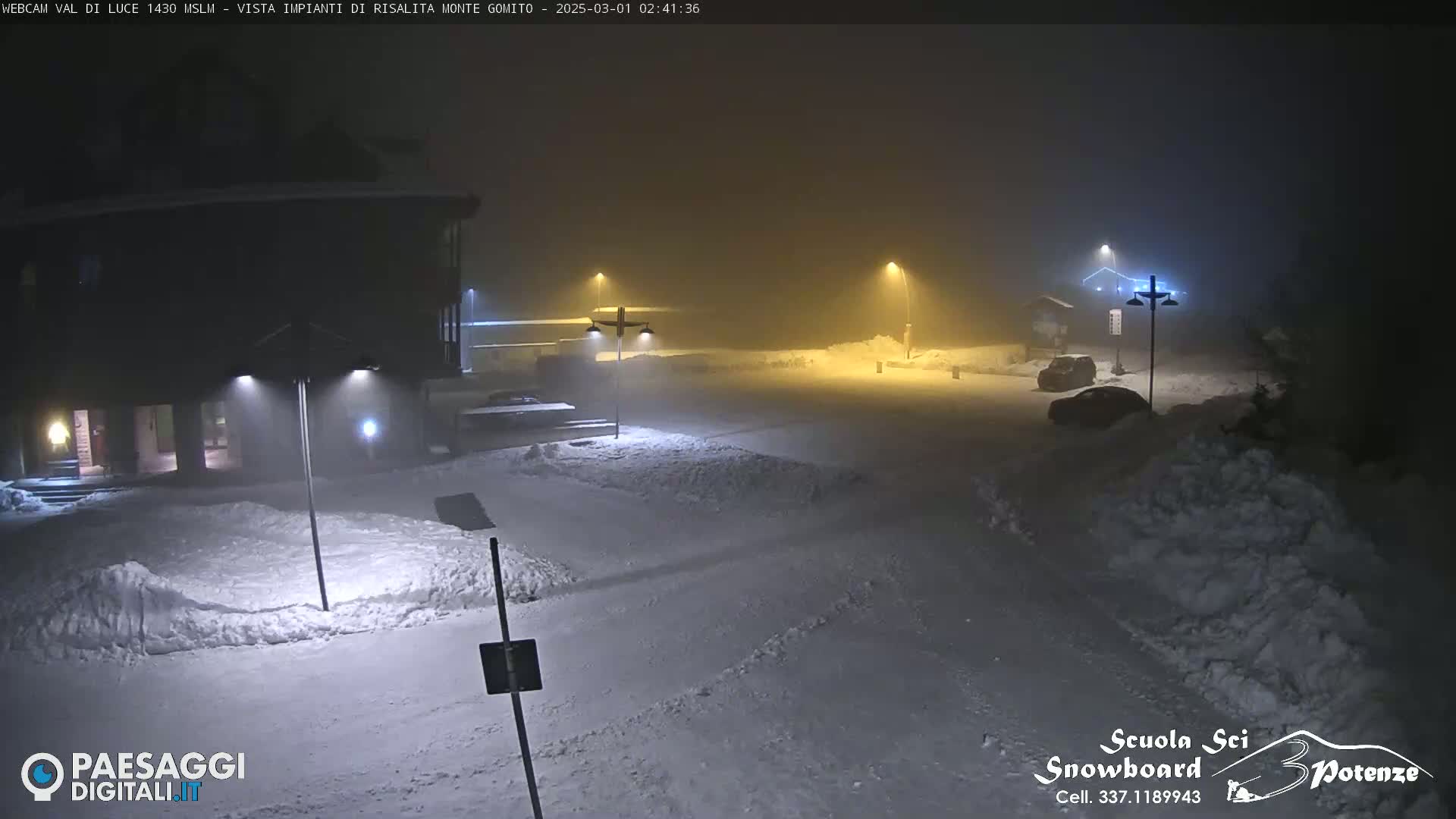 A snow-covered parking lot and building are dimly lit by streetlights in foggy nighttime conditions.
