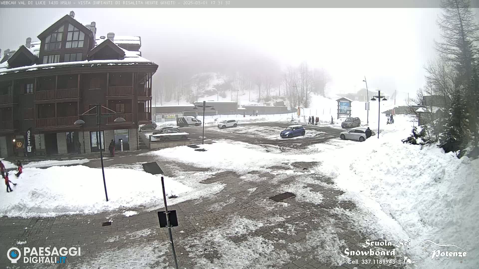 A snow-covered parking lot and plaza sit in front of a multi-story building, all under a foggy, snowy sky.
