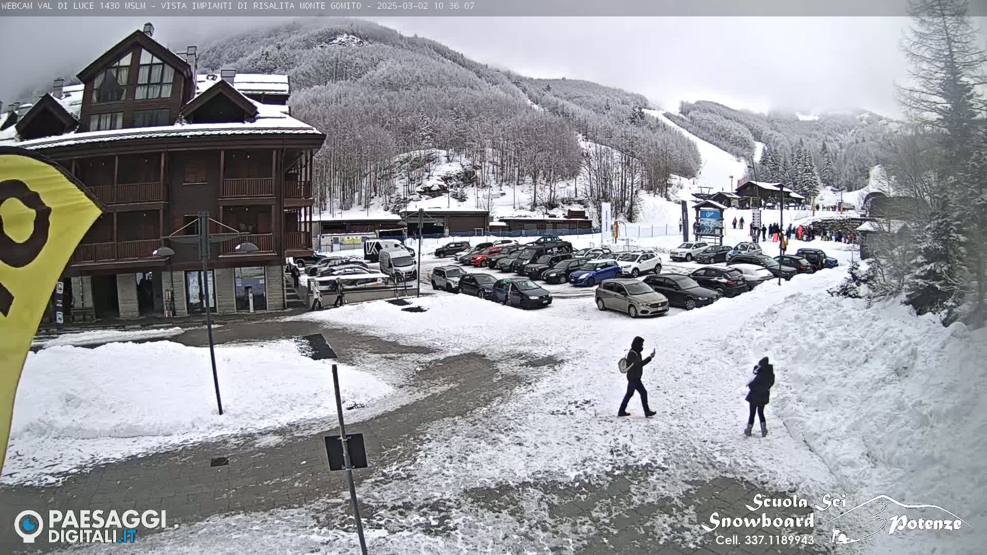 A snow-covered parking lot and plaza are in front of a multi-story building, with a ski slope visible in the background under an overcast sky.