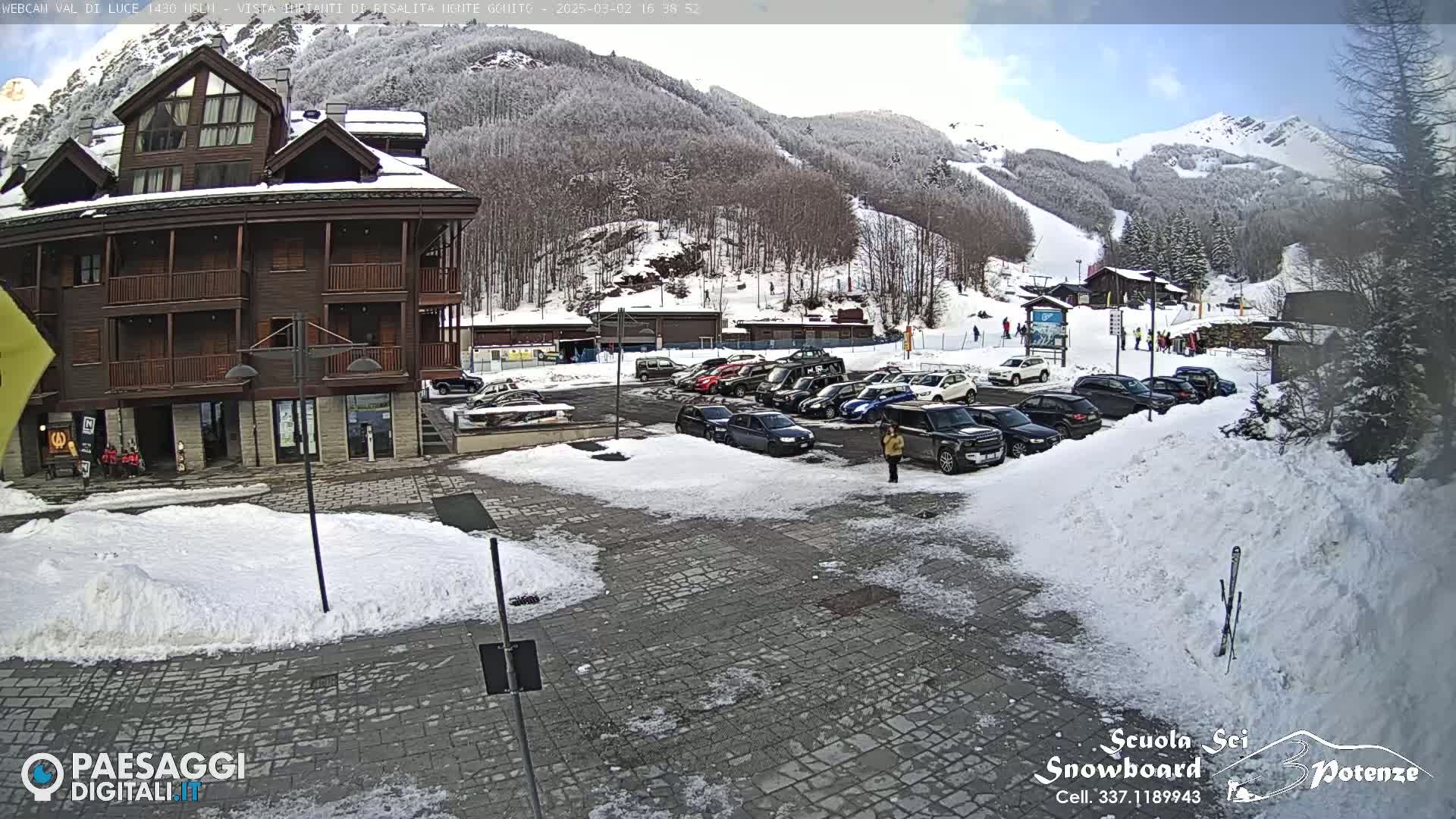 A snow-covered parking lot with cars and a multi-story wooden building sits in front of a snow-dusted mountain with a ski slope.