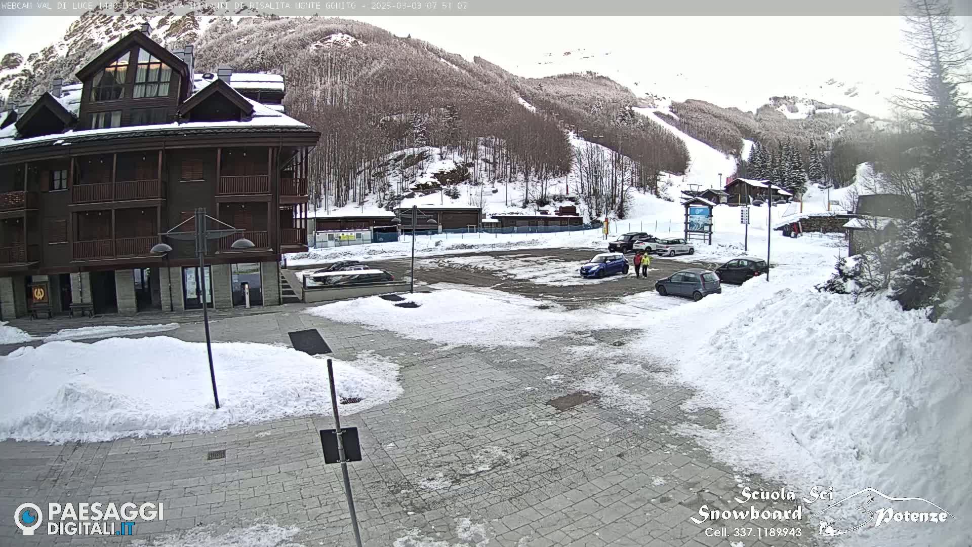 A snow-covered parking lot and paved area are in front of a multi-story building, with a snowy mountainside and ski slope in the background under an overcast sky.