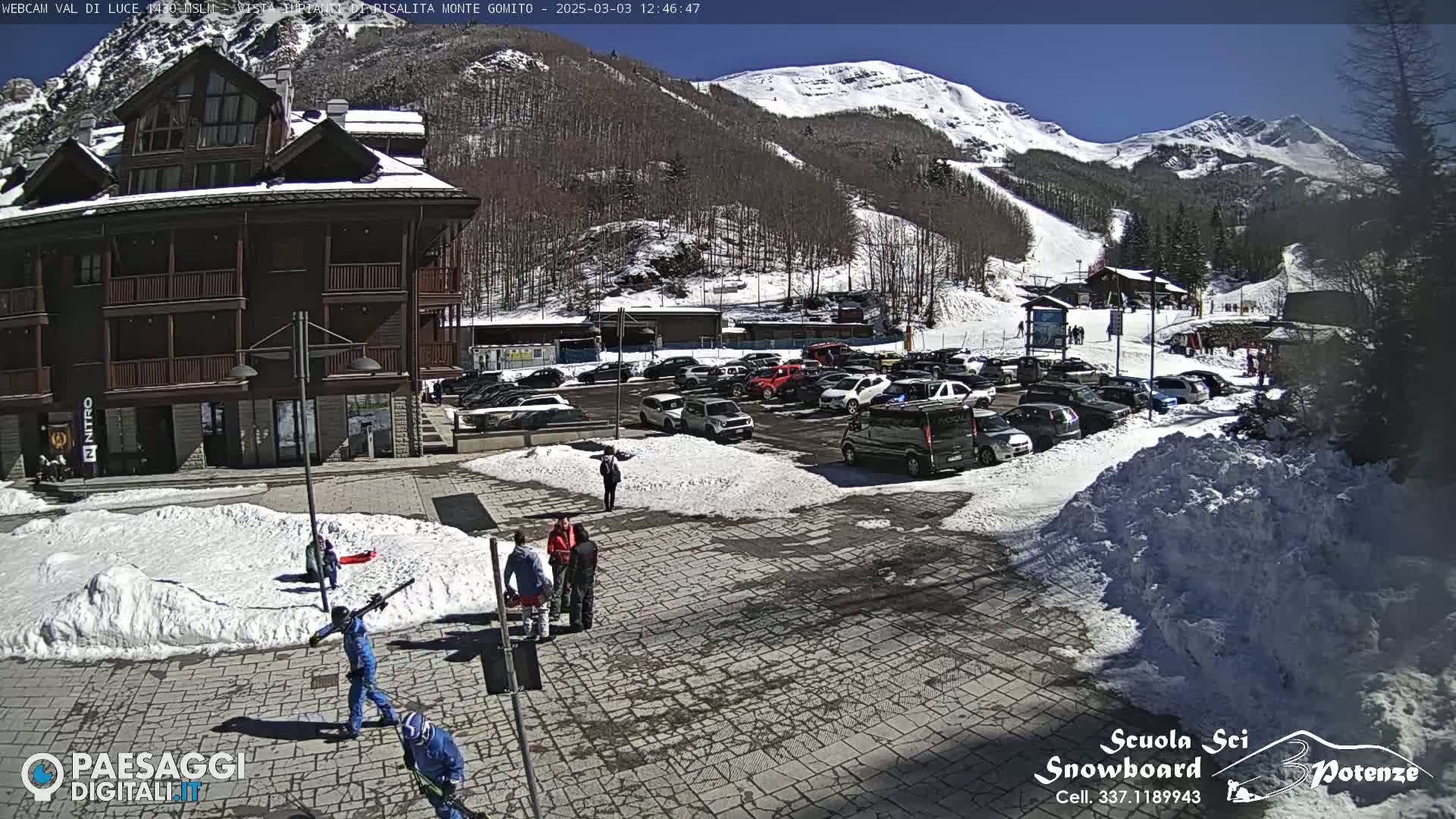 A sunny day at a ski resort shows a parking lot full of cars near a multi-story building, with several people walking around in a snow-covered plaza.