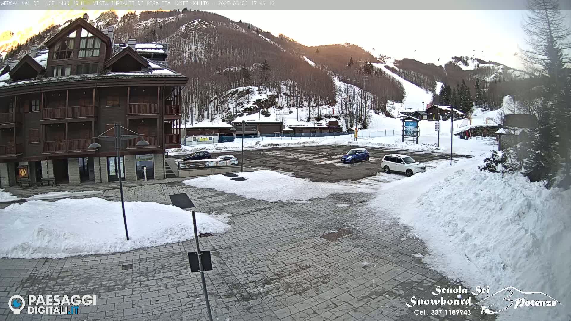 A snow-covered parking lot sits in front of a brown multi-story building, with a snowy mountain and ski slope in the background under a partly sunny sky.