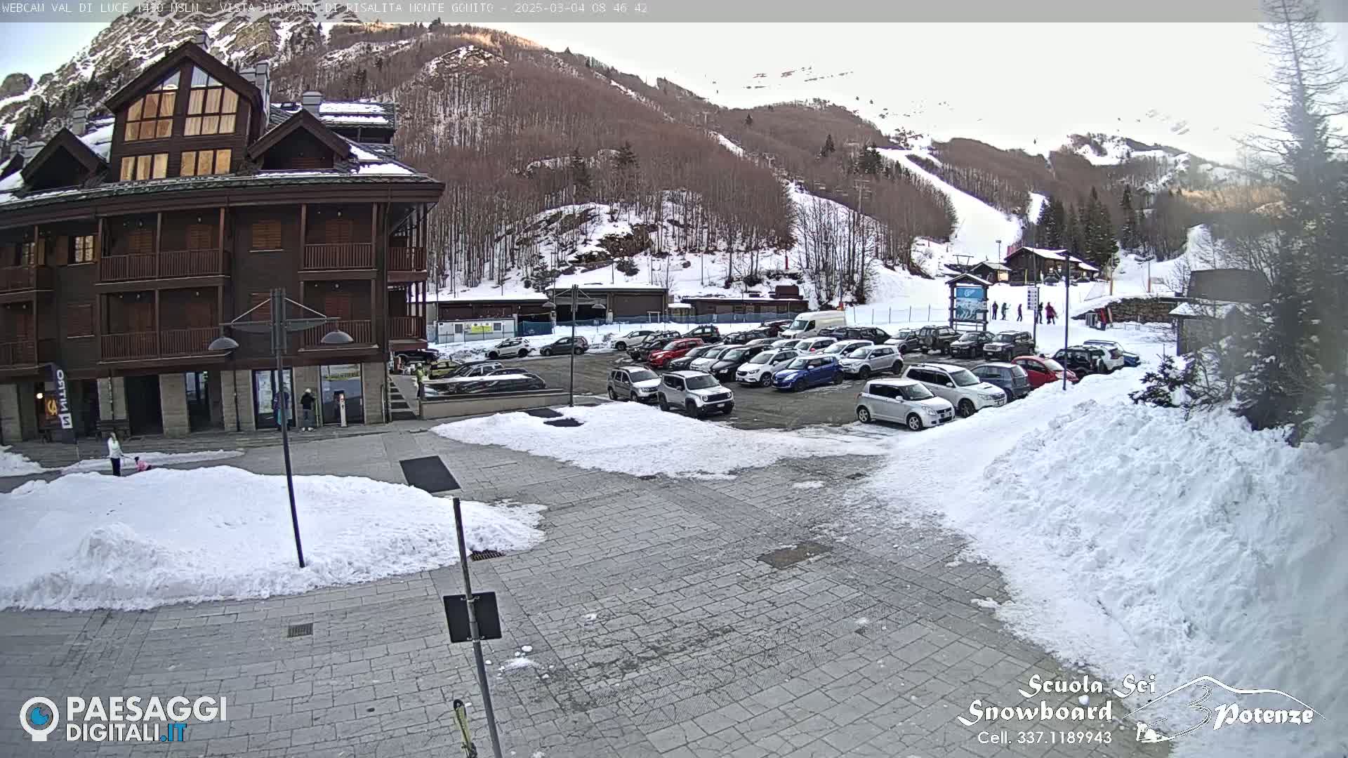 A snow-covered parking lot with cars is in front of a brown multi-story building, with a ski slope visible in the background on a cloudy day.
