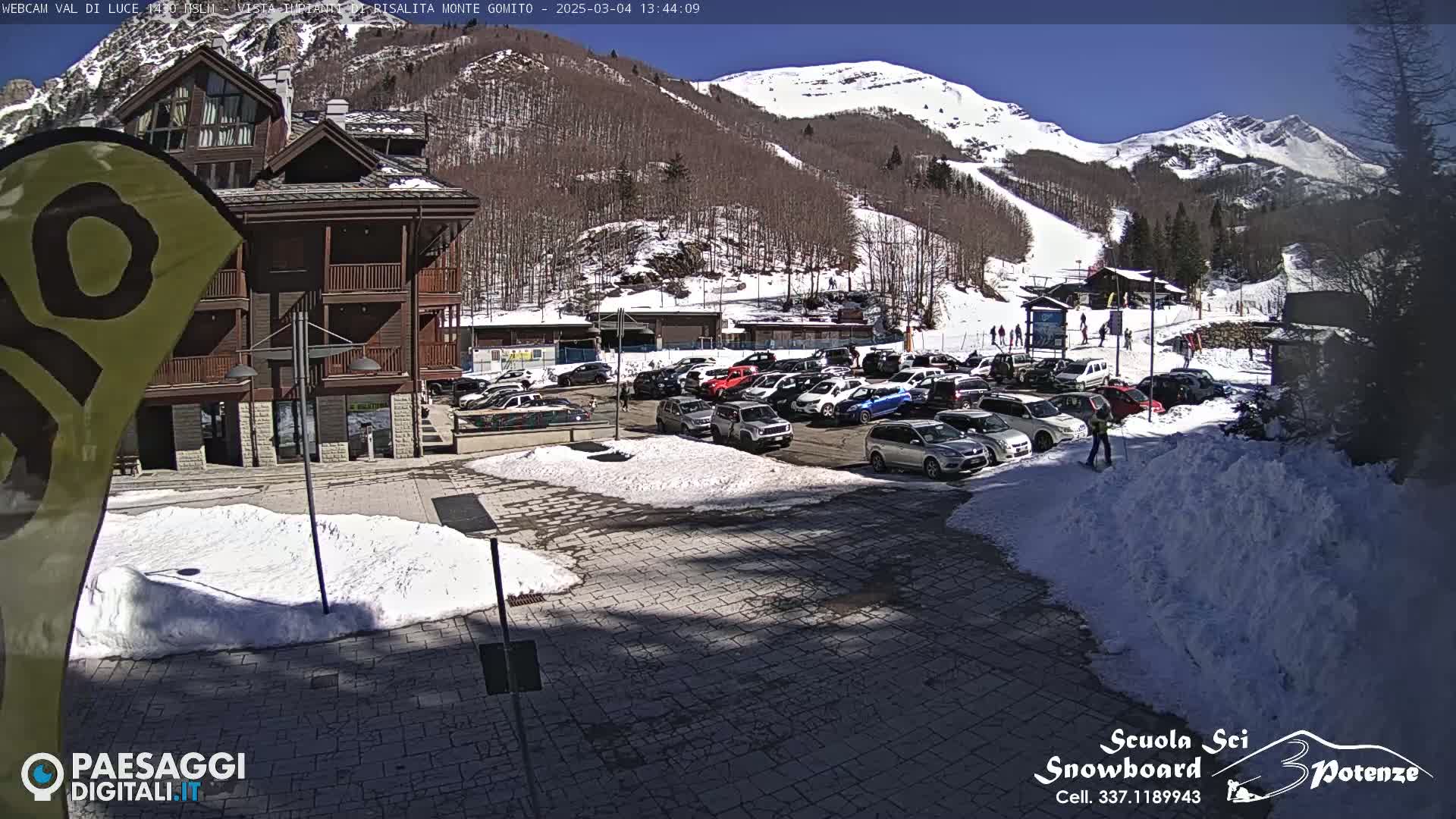 A sunny day at a ski resort shows a parking lot full of cars near a building and ski slope with patches of snow on the ground.