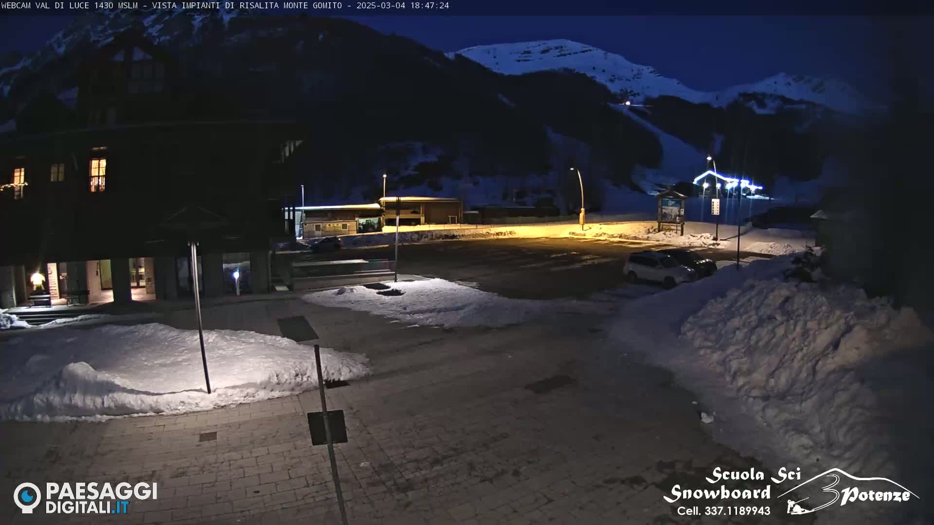A nighttime view of a snow-covered parking lot and building complex, illuminated by artificial lights, with a dark mountainside in the background.