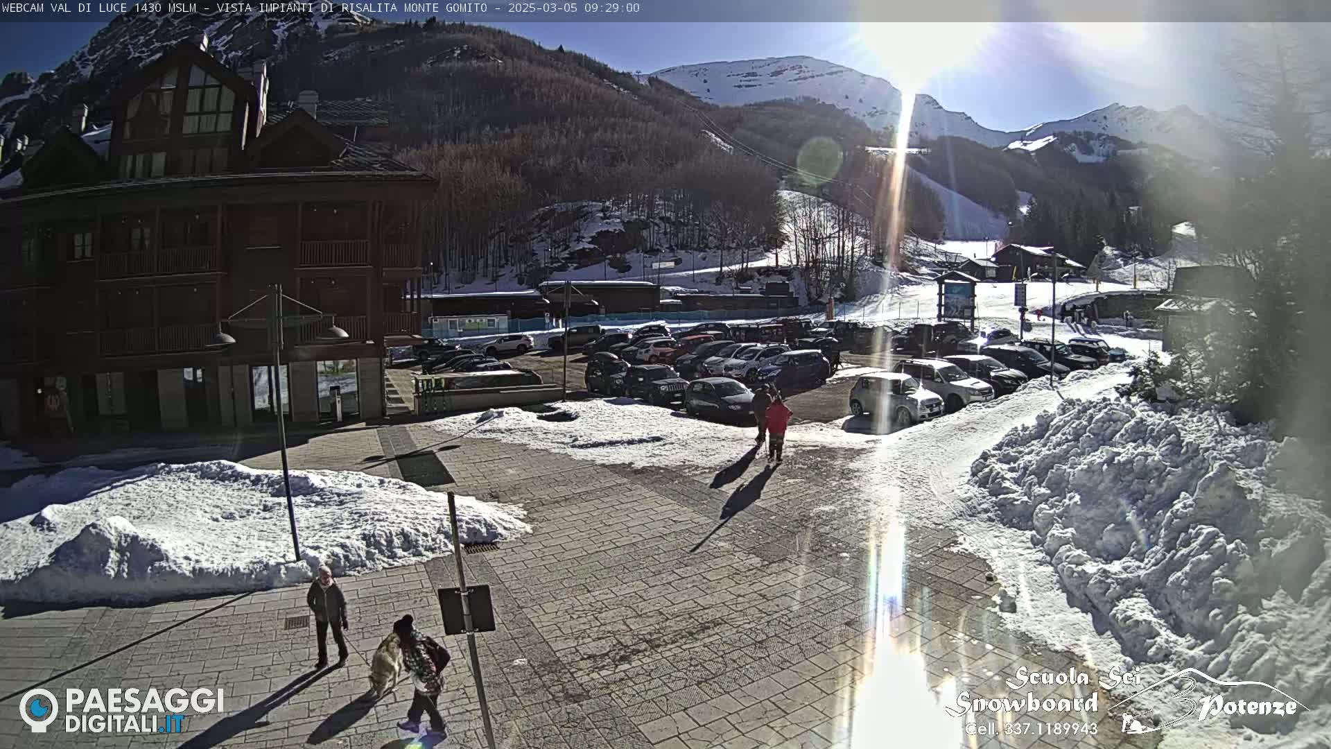 A sunny, snow-covered parking lot with a multi-story building and ski slopes in the background.