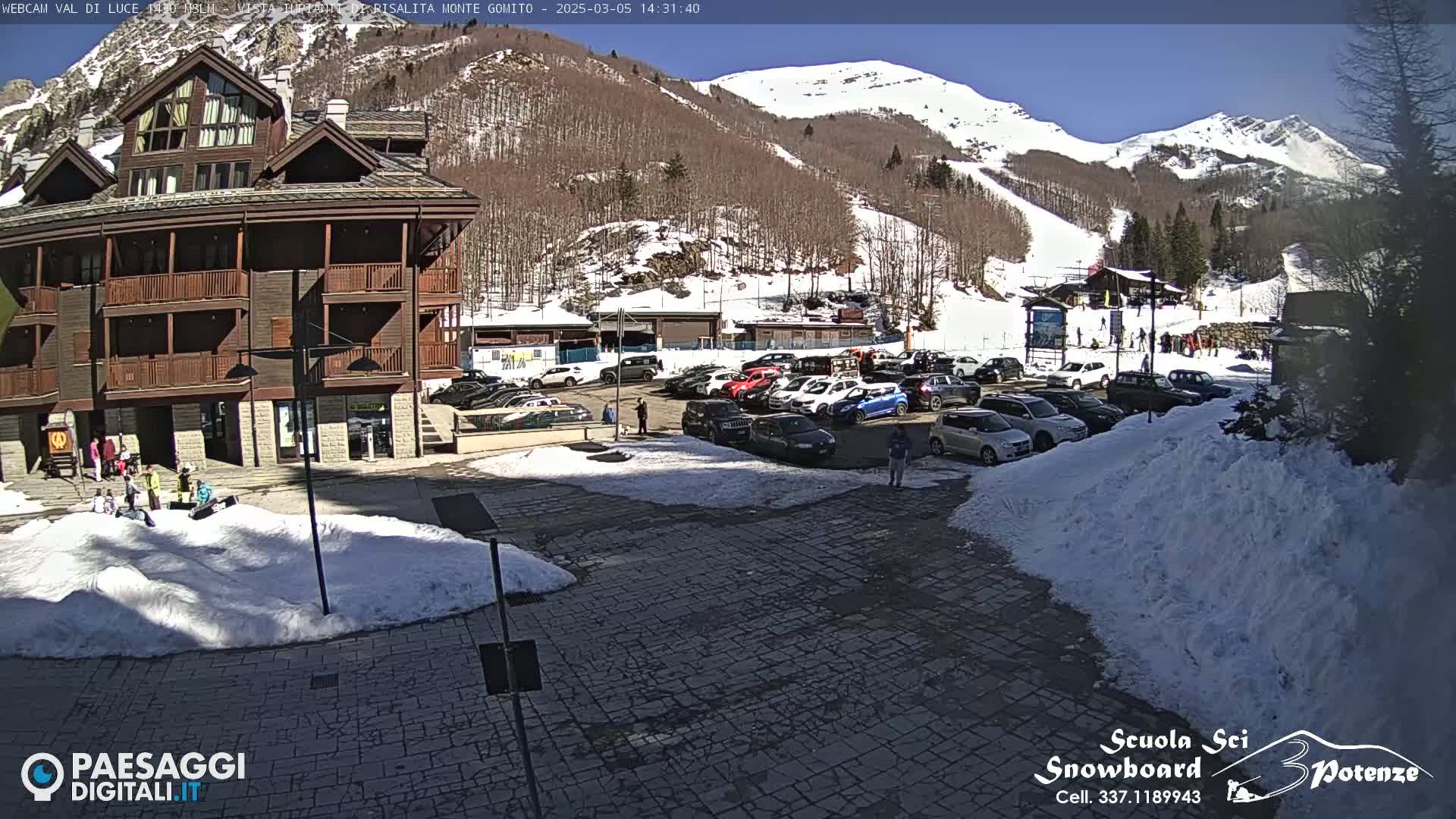 A sunny day at a snow-covered ski resort shows a parking lot full of cars near a multi-story building, with a mountainside ski slope in the background.
