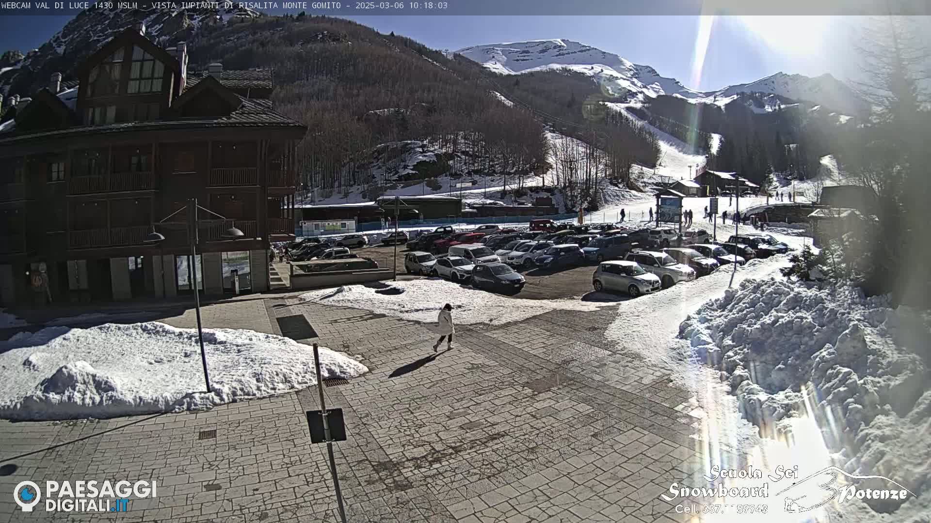 A sunny day at a snow-covered mountain resort shows a parking lot full of cars, a multi-story building, and a person walking on a stone paved area with patches of snow.