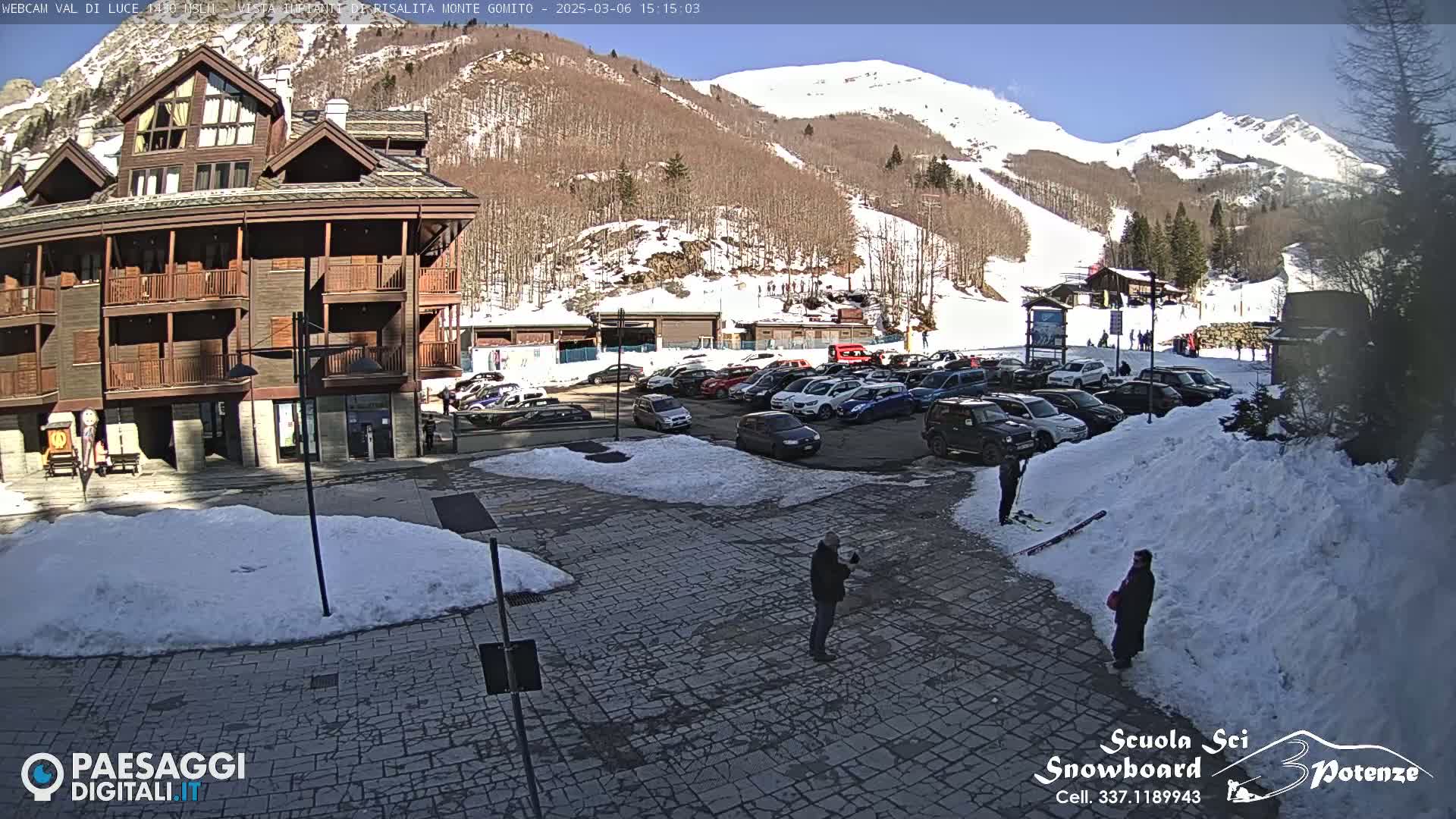 A sunny day in a snow-covered mountain town shows a parking lot full of cars, a multi-story building, and several people standing in a paved area with snow piles nearby.