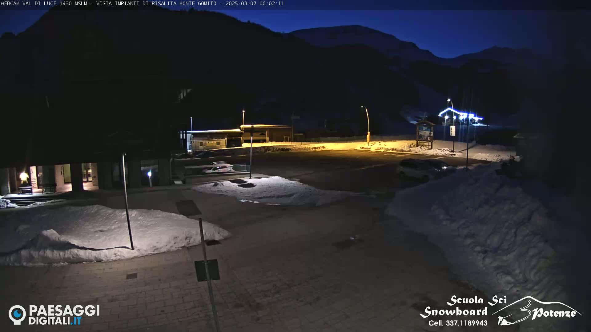 A mostly empty, snow-covered parking lot is illuminated at night by streetlights, with buildings and mountains visible in the background under a dark, clear sky.