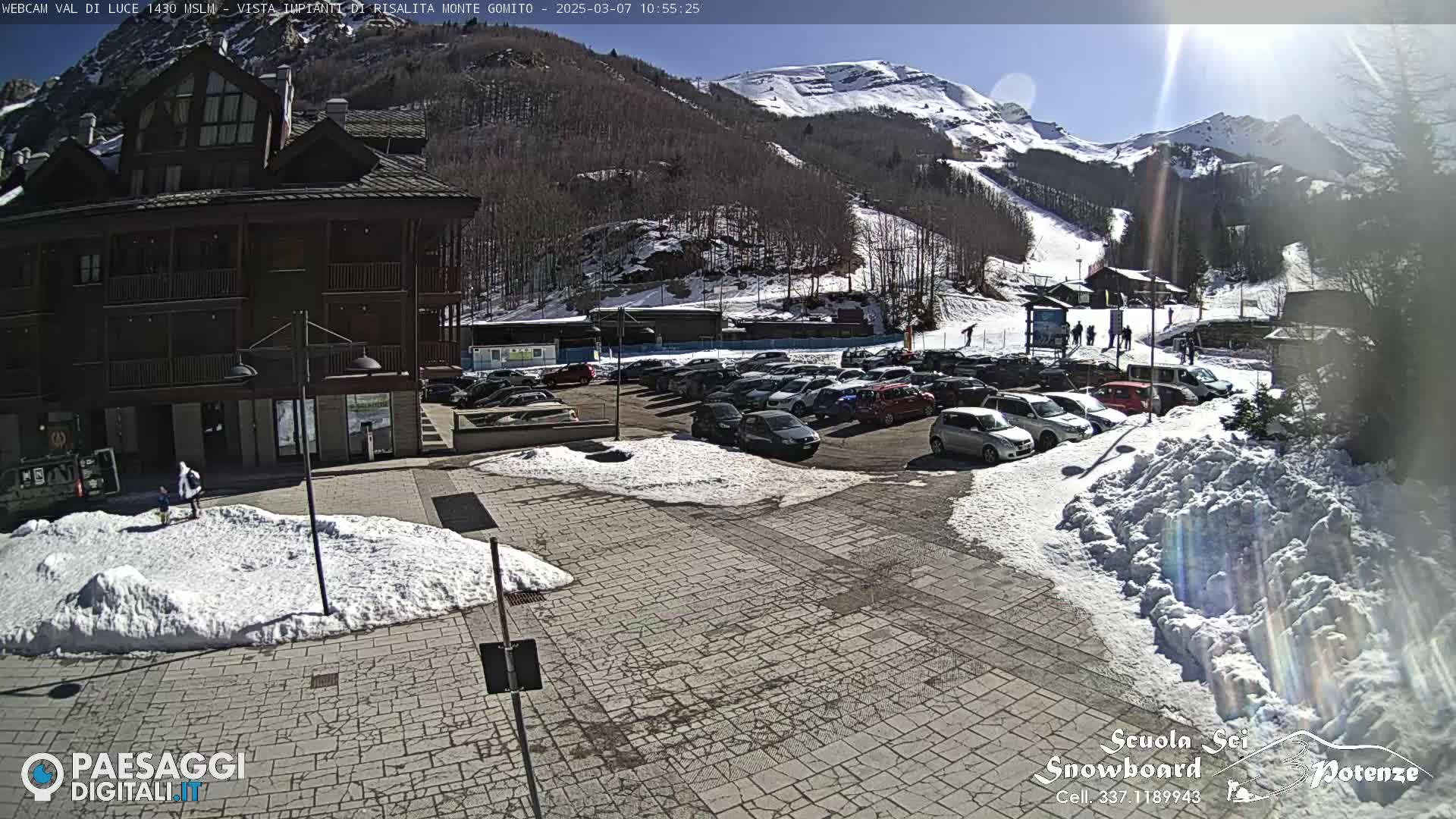 A sunny day at a ski resort shows a parking lot full of cars in front of a brown building, with snow on the ground and a snow-covered mountain in the background.